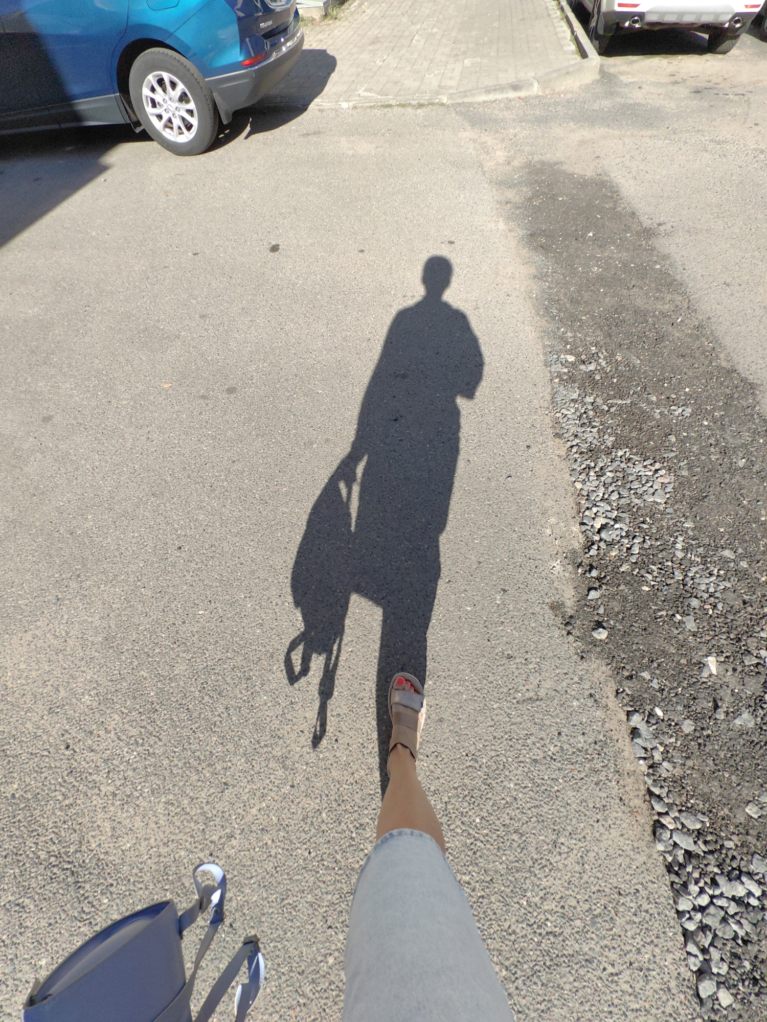 An individual's shadow is cast on an asphalt road, suggesting a sunny day. The shadow depicts a person carrying a backpack. In the foreground, the lower legs and feet of the same individual are visible, wearing a denim skirt and sandals. A blue bag rests on the ground near their feet. To the left, the rear wheel and lower body of a blue car are visible, parked on the side of the road. The background shows a paved sidewalk and part of another vehicle. The scene appears to be a street or parking area.