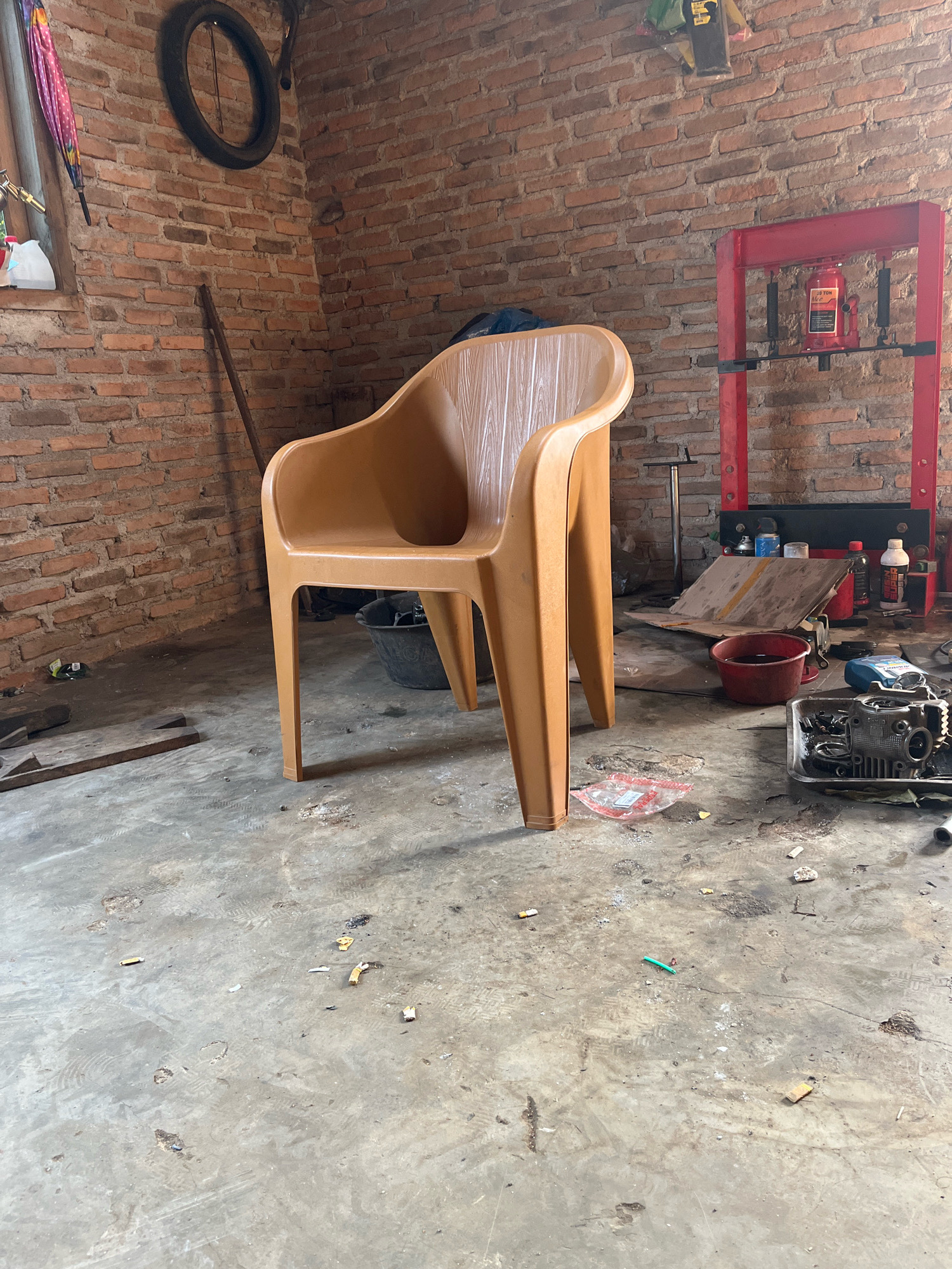 The image depicts a cluttered garage or workshop in Kebumen, Indonesia. The primary focus is a light brown plastic chair positioned in the center of the frame, facing towards the right. The walls are constructed of exposed red brick, and the floor is a concrete surface that is stained and littered with debris, including cigarette butts and what appear to be footprints.
Various tools and automotive parts are scattered around the space. To the left of the chair, a bicycle wheel is mounted on the wall. In the background, a red hydraulic press is visible, with a red jug and various bottles on a shelf. To the right of the chair, a metal tray holds disassembled engine parts. Other items present include a black bucket, a broom leaning against the wall, and a crumpled plastic bag on the floor. The lighting suggests an indoor environment, possibly with natural light from a window or overhead lighting, casting shadows on the floor. The overall impression is of an active but untidy workspace.