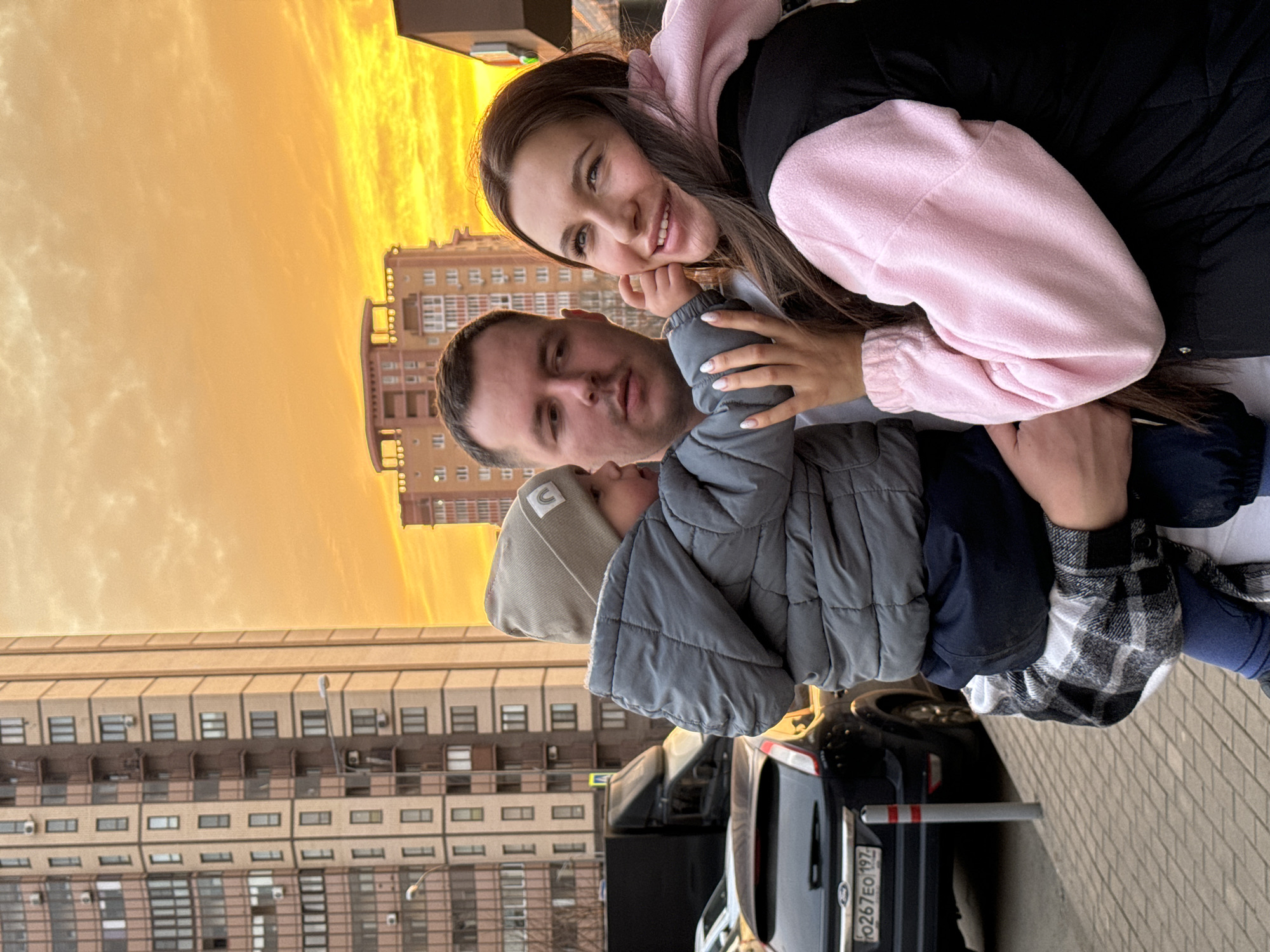 A mid-shot photograph captures three individuals—two adults and one child—standing on a paved sidewalk in an urban setting in Moscow, Russia, identified by the Russian license plate "O267EO 197" on a visible vehicle. The sky in the background is a vibrant orange and yellow, indicative of either a strong sunset or sunrise.
On the left, a male adult with short dark hair wears a white t-shirt beneath a black and white plaid shirt. He holds a young child in his arms, facing the child towards the right. The child wears a grey puffer jacket, dark blue trousers, and a light grey beanie adorned with a small rainbow emblem. The child's left hand is placed on the cheek/ear area of the female adult to their right.
The female adult, positioned on the right side of the frame, has long dark hair and wears a black outer garment over a pink fleece hoodie. She is smiling and looking upwards and to her right.
In the background, multiple multi-story residential buildings are visible, characterized by light brown and tan facades with numerous windows and balconies. A black Ford Focus hatchback is parked behind the subjects, displaying the Russian license plate "O267EO 197." Further back, a dark-colored truck is also present. A grey and orange-red striped bollard is visible on the sidewalk near the parked Ford.