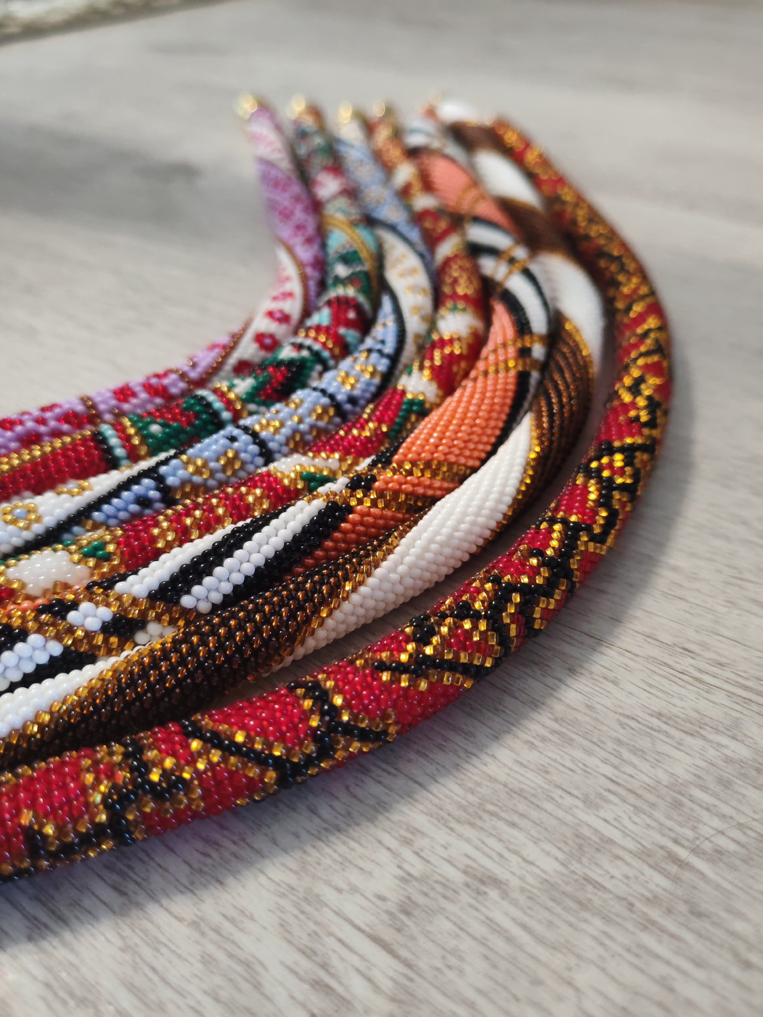 A close-up, overhead shot displays a collection of beaded necklaces arranged in a curved fashion on a light-colored, wooden surface. The necklaces are intricately designed with various colorful bead patterns, including red, white, blue, green, purple, orange, black, and gold. The beads appear to be small and tightly woven together, creating textured, cylindrical forms. The lighting is soft, casting subtle shadows and highlighting the reflective quality of some of the beads. The background is out of focus, with a slight blur that emphasizes the foreground subjects. There are no people or animals visible in the image. The overall impression is one of craftsmanship and decorative artistry. The location is Unknown, Unknown.