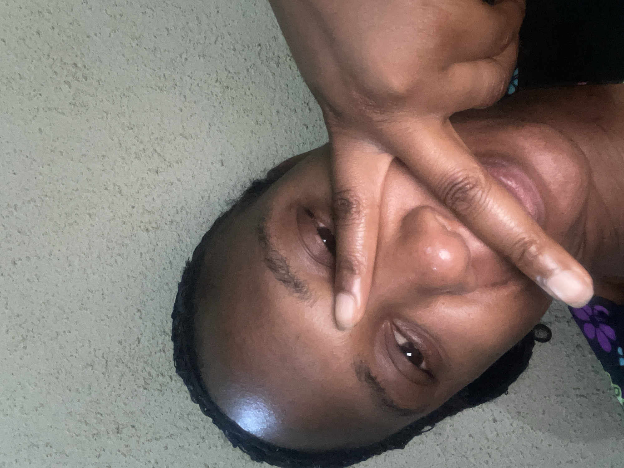 A close-up, slightly rotated, image shows a dark-skinned individual with short, braided hair. The person is holding their hand up, obscuring part of their face, specifically covering one eye and a portion of their forehead and nose. Only one eye is visible. The background appears to be a textured wall, possibly painted in a light, neutral color. Due to lack of information, it is impossible to provide details on the location of Jos, Nigeria.