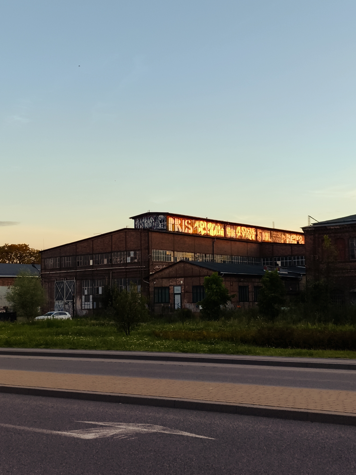 An industrial building with graffiti across its top facade is visible against a clear blue sky with a hint of orange from the setting sun. The building, constructed of red brick, features large windows, many of which appear broken or boarded up. A white car is parked in the grassy area in front of the building, which also has several trees. A road with a pedestrian walkway runs in the foreground of the image. The graffiti on the building appears to read "PRISMA". This is an image of the former "Pryzmat" factory in Gdańsk, Poland.