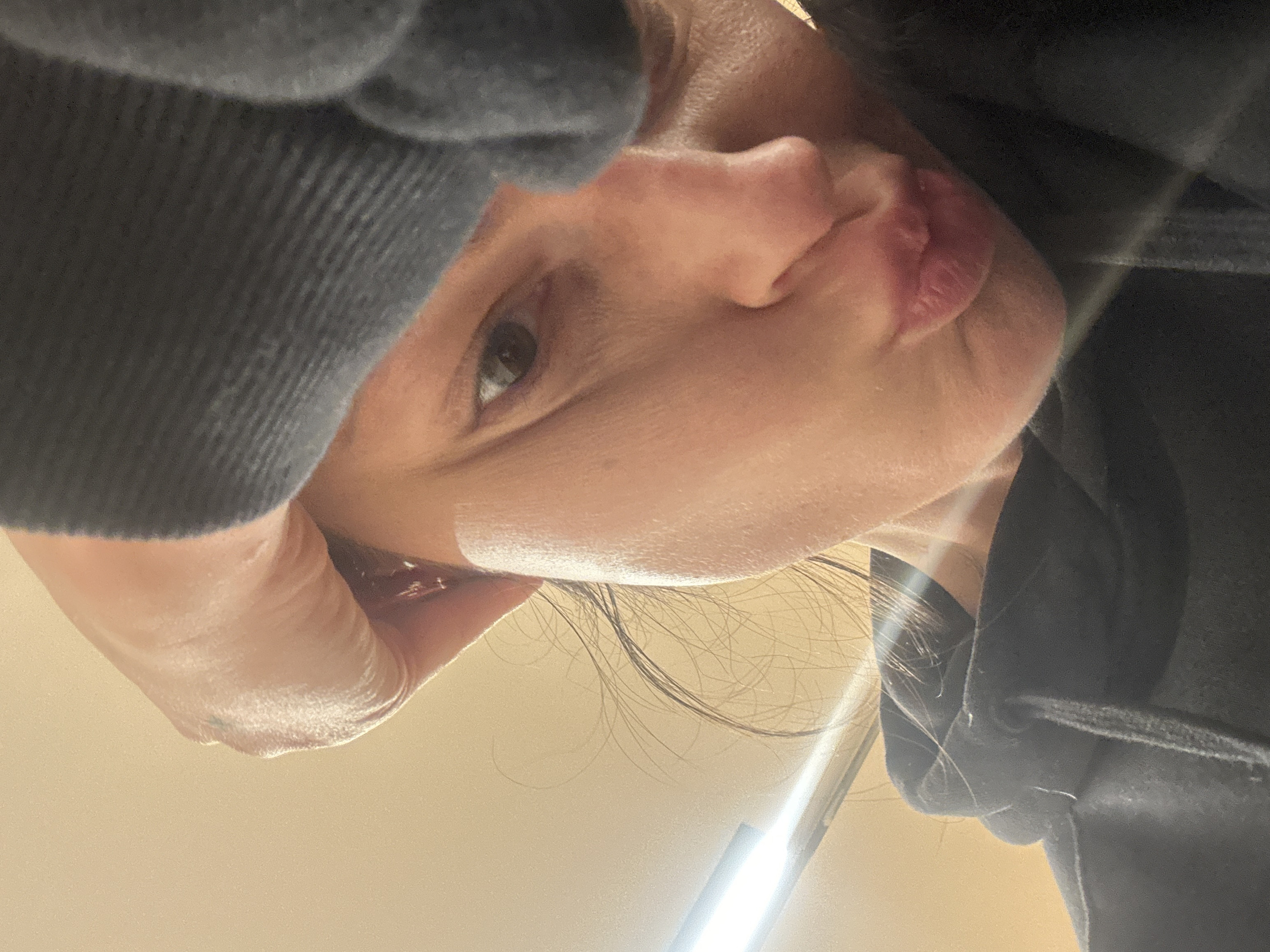 The image presents a close-up, low-angle perspective of a person's face, tilted downwards and to the right, looking up towards the viewer. The person is wearing a dark, possibly charcoal grey or black, hooded garment, likely a hoodie or sweatshirt, with a visible ribbed texture on the fabric, particularly on the left sleeve which partially covers the forehead.
The visible features of the face include one eye, which appears to be the left eye (from the viewer's perspective), looking directly at the camera. The iris is dark brown or black, and a bright, rectangular reflection of a light source is clearly visible within the pupil. The bridge and tip of the nose are prominent, showing natural skin texture and slight redness. The upper lip is visible, appearing full and slightly pursed or compressed, with some natural lines. The skin complexion is fair to medium, with visible pores and a subtle sheen.
Dark hair strands are visible framing the face, particularly on the right side and falling towards the bottom of the frame. A strong, artificial light source, likely a fluorescent tube or LED strip, is visible as a bright, elongated streak in the bottom right corner of the image, illuminating parts of the face and creating the reflection in the eye.
The background is a plain, light-colored surface, possibly a wall or ceiling, suggesting an indoor setting. The overall composition gives the impression of a candid or self-taken photograph, capturing a moment of direct gaze. No text is visible in the image.