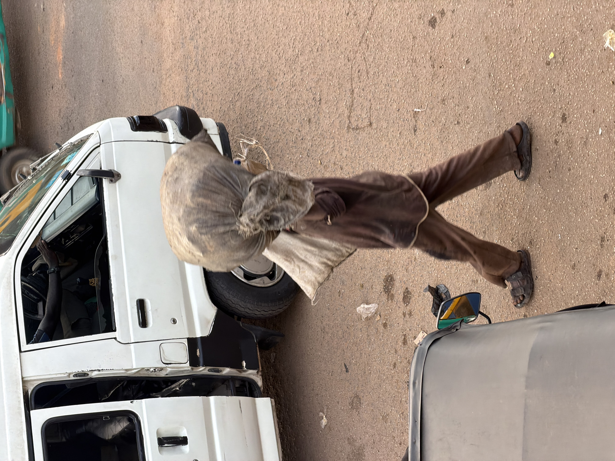 In Bukuru, Nigeria, a person walks on a dirt road carrying a large, burlap-wrapped bundle on their head. The person is wearing brown pants and a brown shirt, and dark-colored sandals. To the left, a white van with its side door open is parked on the side of the road. The van has black trim around the windows and on the lower body. In the foreground, the edge of a dark-colored vehicle, possibly a tuk-tuk, is visible. The ground is a textured, reddish-brown dirt. Scattered on the ground are pieces of white plastic trash. The image is taken from a slightly elevated perspective, looking down on the scene.