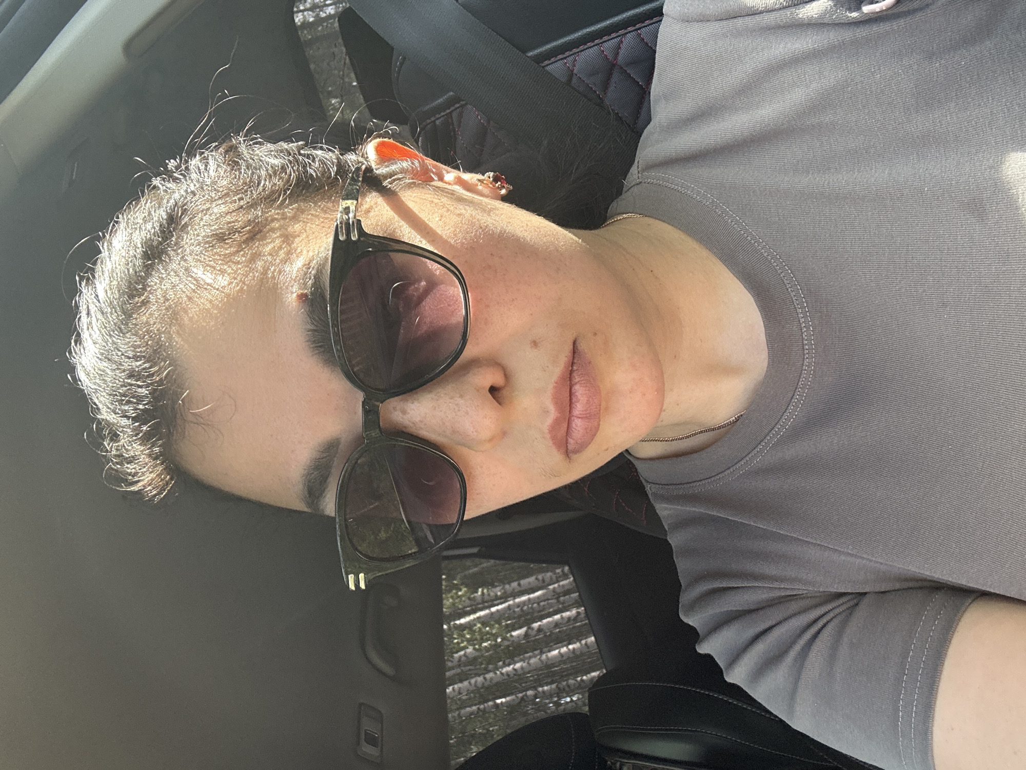 A person wearing sunglasses and a grey shirt is sitting in a car. The location appears to be within the city of Unknown, Unknown. The interior of the car is visible, including a seatbelt and a headrest. Trees are visible through the car window.