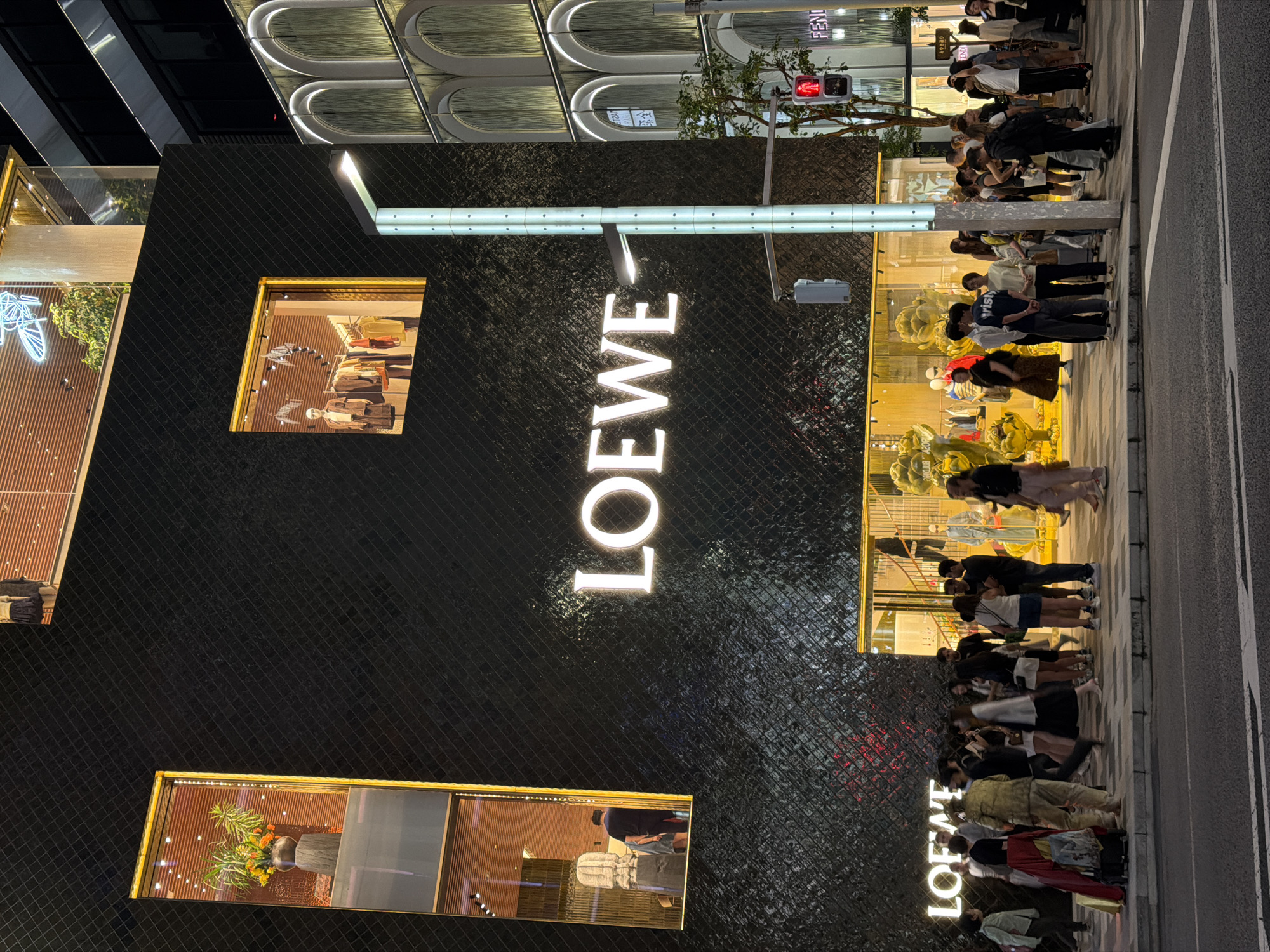The image captures a street scene at night in front of the Loewe store in Chuo, Japan. The building's facade is dark and textured, featuring a large, illuminated "LOEWE" logo. There are two prominent windows on the upper floors, one displaying a vase with flowers and the other showing clothing on racks with a mannequin. A busy sidewalk is in front of the store, populated by a crowd of people, many of whom are standing and looking towards the store or talking to each other. The ground floor of the store has large glass windows revealing displays with mannequins and decorative elements. To the right of the Loewe store, another building with an arched facade and illuminated windows is visible. A traffic light with a red pedestrian signal is present on the right side of the image, and a street lamp illuminates the sidewalk. The street in the foreground is paved, and a crosswalk is marked. The overall atmosphere is one of a bustling urban environment at night, with a focus on a luxury retail establishment.