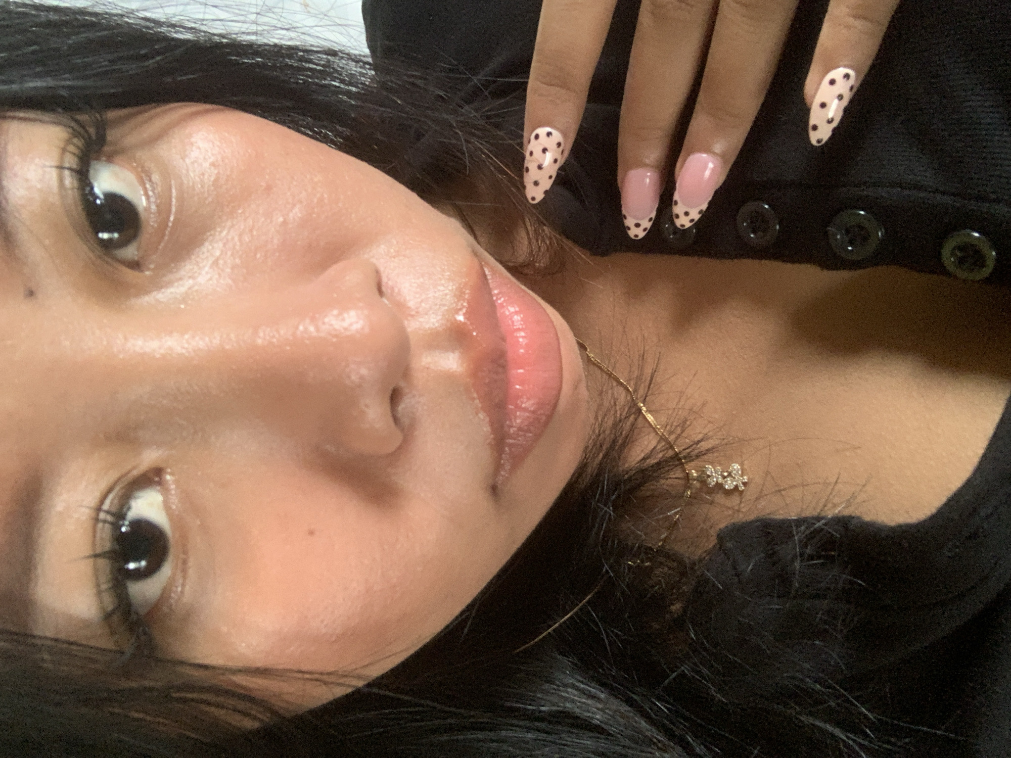 A close-up, low-angle selfie shows a person's face and hand against a black garment. The person has dark hair, dark eyes with long eyelashes, and light-pink, glossy lips. Their skin appears to have a dewy sheen. The person is wearing a gold necklace with a small, diamond-encrusted charm. Their fingernails are decorated with pink French tips and black polka dots. The person's right hand is positioned near their face, with fingers slightly curled. The background is nondescript and appears to be an indoor setting. There is no visible indication of the city of Jajag, Indonesia, or any specific environmental features that would place it there.