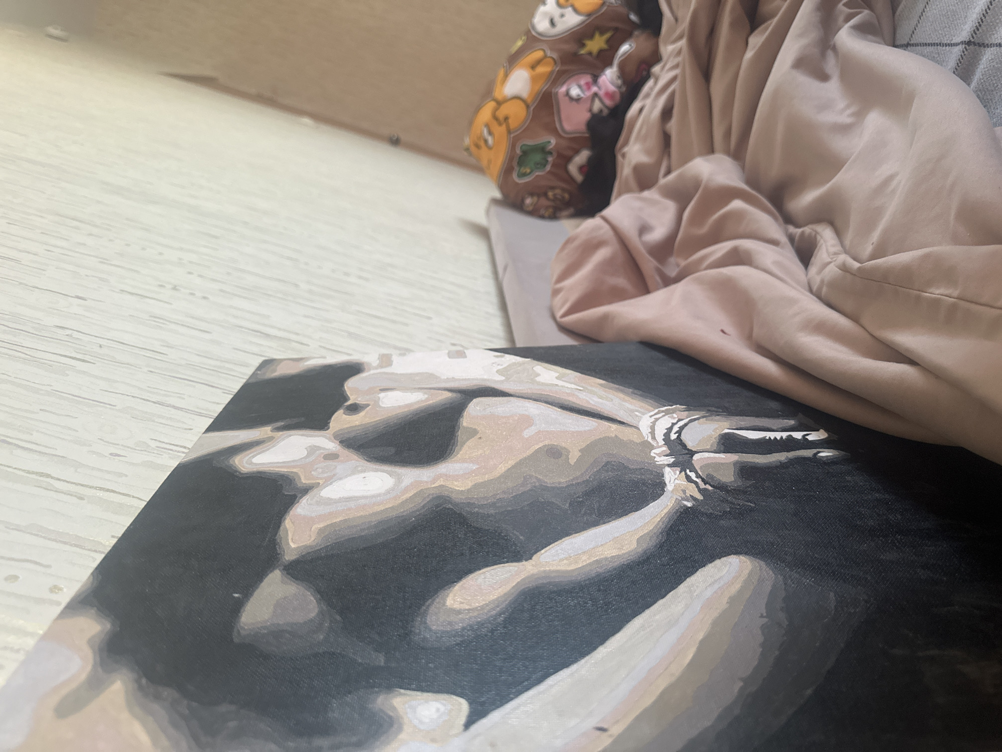 The image shows a painting of a nude figure rendered in grayscale tones, with the figure appearing to be bound or constrained. The painting is resting on a light-colored, possibly wooden surface, likely a table or floor. In the background, there's a rumpled light brown fabric or garment and a cushion or pillow with cartoon-like characters on it. The setting seems to be indoors, possibly a bedroom or living area. The location is Высокий, Russia.