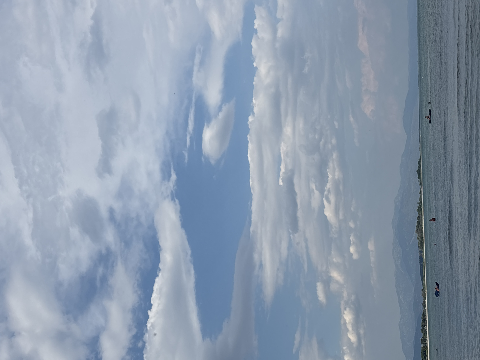 This media file captures a picturesque scene by the sea, likely in Mandra, Greece. The image is oriented vertically, with the sky dominating the left half and the sea and distant shoreline occupying the right.
The sky is a canvas of dramatic clouds, ranging from wispy white formations to denser, darker masses, suggesting a dynamic weather pattern or possibly the transition towards evening. Patches of bright blue sky peek through, indicating daylight.
The sea on the right is calm, with gentle ripples reflecting the muted light from the sky. Along the distant horizon, a line of mountains forms the backdrop, with a strip of green trees at their base, hinting at a coastal landscape. The overall mood is serene and tranquil, with a sense of natural beauty.
Several figures are present in the water. In the middle distance, a person is visible on a dark paddleboard. Further out, another individual appears to be swimming or standing in the water. Towards the far right, a person in a red top is visible in the water. These figures suggest recreational activities are taking place, possibly swimming, paddleboarding, or simply enjoying the water.
The time of day appears to be late afternoon or early evening, given the soft, diffused light and the hues in the sky and on the water. The weather seems pleasant, with a mix of sun and clouds. No text is visible in the image.