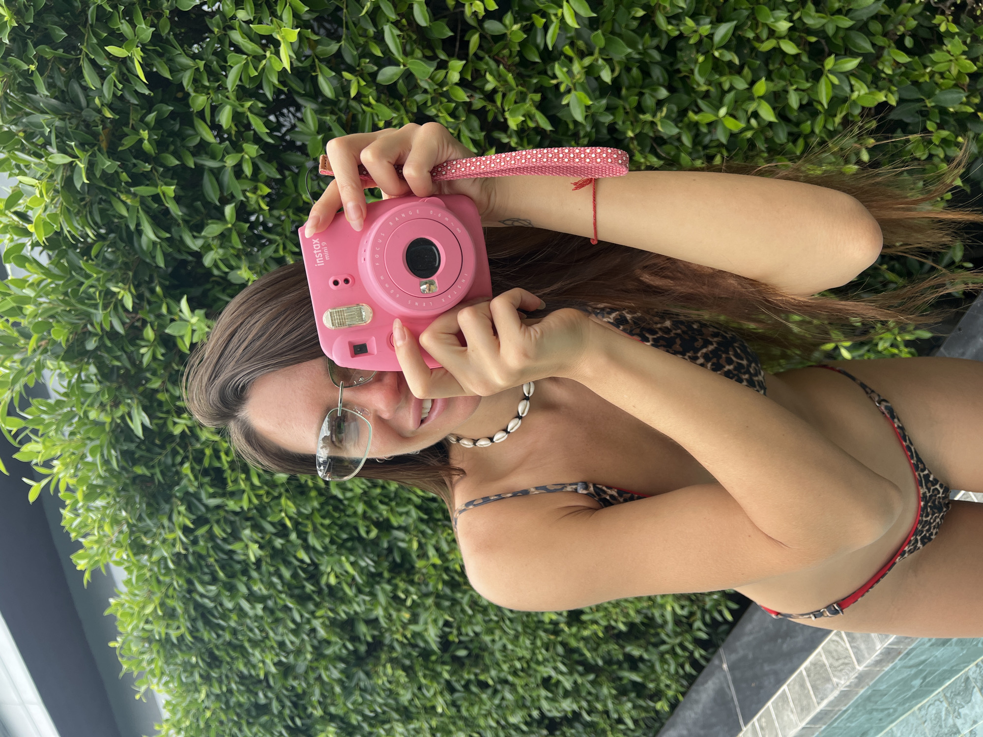 A young woman is depicted in a cheerful outdoor setting, likely a poolside or similar water body in บ้านบางรักษ์, Thailand. She is smiling broadly and holding a bright pink Instax mini 9 instant camera, positioned as if ready to take a picture. Her long brown hair frames her face, and she is wearing stylish clear-framed sunglasses and a white shell necklace.
She is dressed in a leopard print bikini with subtle red detailing, and a thin red string bracelet is visible on her right wrist. The pink camera features a white polka dot strap. The upper half of her body is visible, with her arms raised to hold the camera. The background is filled with lush, vibrant green foliage, suggesting a tropical or well-maintained garden environment. The foreground shows the tiled edge of the water body she is partially in. The bright, natural lighting indicates it is daytime, and her happy expression conveys a relaxed and joyful mood. The camera clearly displays the text "Instax mini 9".