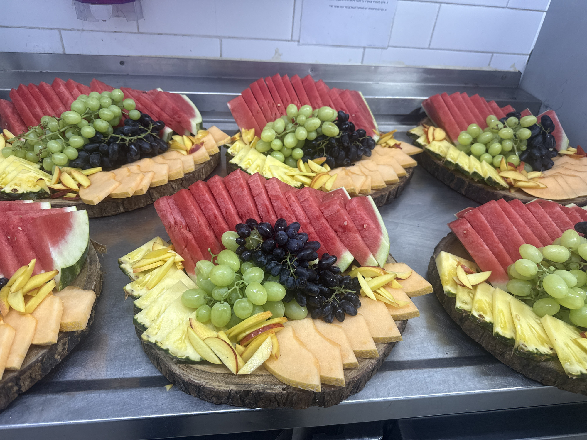 The image shows a close-up, eye-level view of a buffet table in a food establishment, likely in Herzliya, Israel, given the text on the wall sign which appears to be in Hebrew. The table is made of stainless steel, and there are several wooden platters arranged on it, each filled with a variety of cut fruits.
The platters are oval-shaped and have a rustic look with the bark of the wood still visible around the edges. Each platter is decorated with an assortment of colorful fruits, including sliced watermelon, green and black grapes, sliced cantaloupe, sliced pineapple, and sliced peaches. The fruits are arranged in a visually appealing manner, with the watermelon slices fanned out, grapes clustered in the center, and other fruits forming decorative borders.
The background shows a white tiled wall and some overhead lighting. The overall impression is one of a well-presented and fresh fruit buffet. No people are visible in the image. The scene is static and focuses solely on the food display.