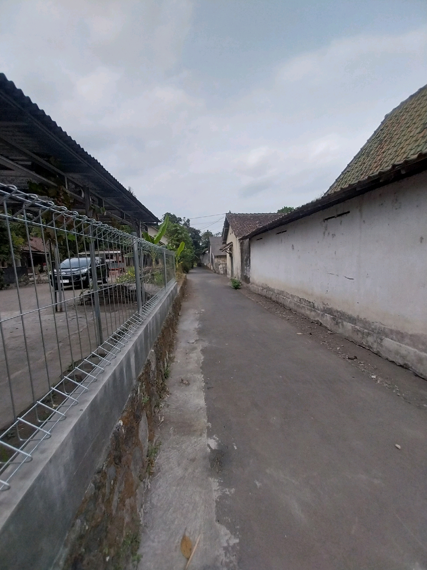 The image depicts a narrow asphalt road in what appears to be a residential area, likely in Tijayan, Indonesia. The sky is overcast, with a muted gray tone. On the left side of the road, a metal fence with a concrete base runs along the edge. Beyond the fence, a dark-colored car is parked in front of a building with a corrugated metal roof, possibly a carport or an open-sided structure. Lush green vegetation is visible behind the fence and on the right side of the road, adding to the rural or suburban feel.
The right side of the road is lined with a long, whitewashed wall, characteristic of many Indonesian homes. The wall shows signs of age, with some discoloration and minor cracks. Further down the road, more buildings with tiled roofs can be seen. The road itself is in fair condition, with some patches of wear and debris scattered along its edges. There are no people visible in the image. The overall impression is a quiet, perhaps slightly run-down, street on an overcast day.