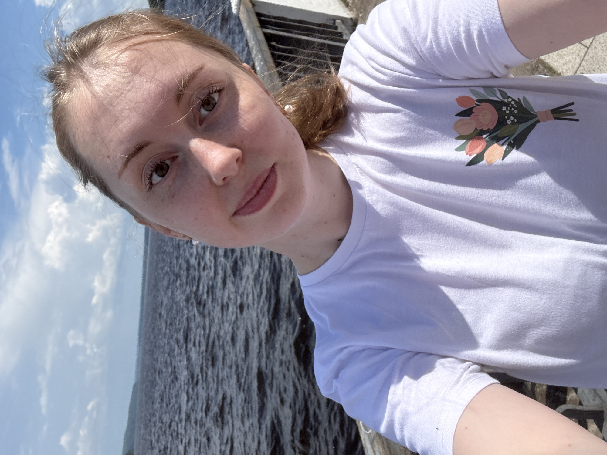 A young Caucasian woman with blonde hair and freckles is taking a selfie outdoors. She is wearing a white t-shirt with a bouquet of flowers printed on the left side of the chest. She has a pearl earring in her right ear. The background shows a body of water and a cloudy sky. The woman is leaning slightly and looking at the camera with a gentle smile. The location appears to be a waterfront promenade or pier.
