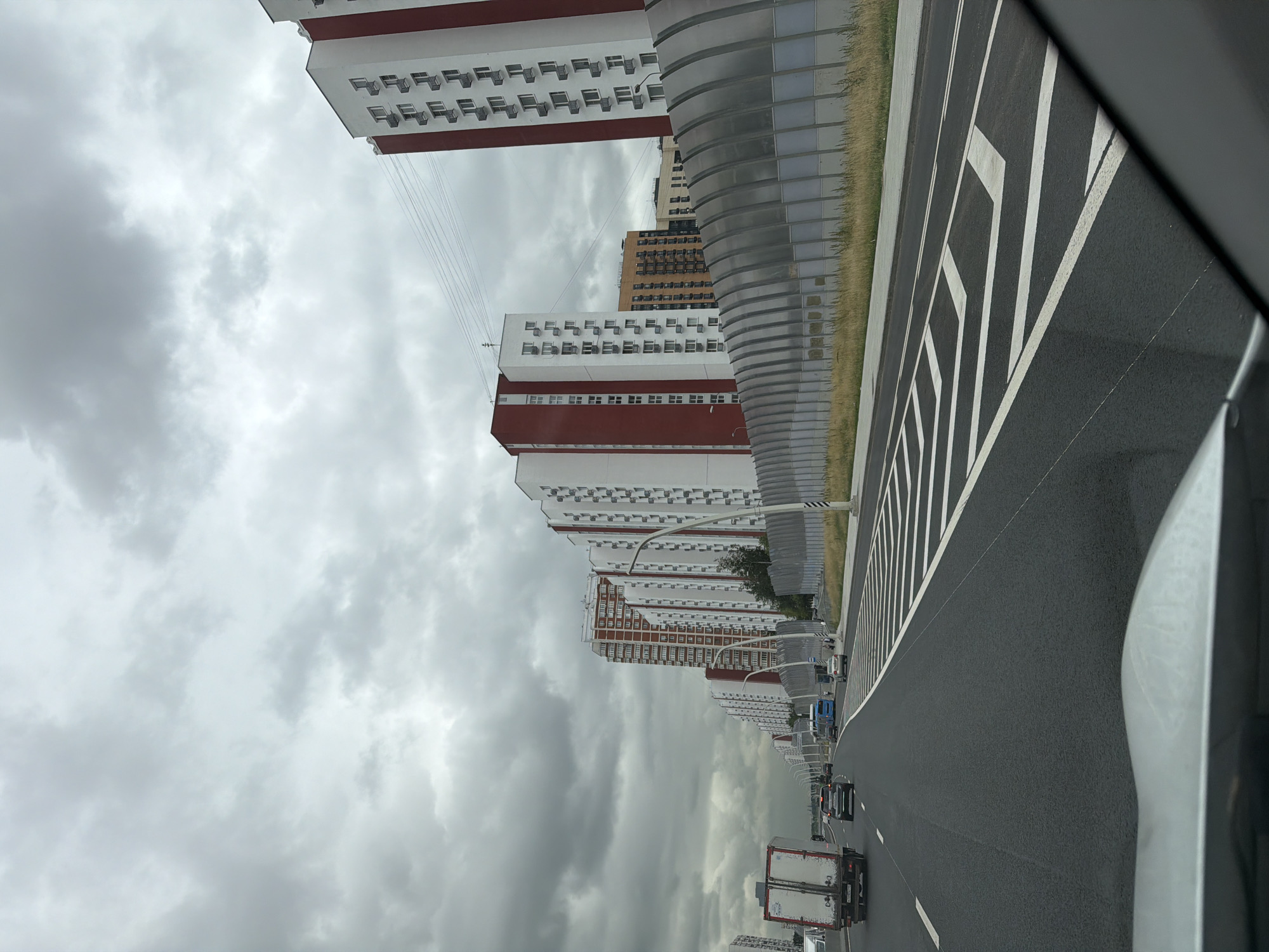 The image depicts a view from a car driving along a modern, multi-lane road. The road is paved with asphalt and marked with white lines. The buildings lining the road are tall, modern residential complexes with distinctive red and white facades, featuring numerous windows and air conditioning units. The architecture appears uniform, suggesting a planned urban development. To the right of the road, there's a section of tall grass and a futuristic-looking, curved metallic structure that seems to be a sound barrier or part of the urban infrastructure.
The sky is overcast with heavy, gray clouds, indicating a cloudy or potentially rainy day. The lighting is diffused, suggesting it is daytime, but without direct sunlight. The image captures a sense of urban transit, with several vehicles visible on the road, including trucks and cars, moving in the same direction as the viewpoint. The overall scene is one of an active, modern urban environment in Moscow, Russia, characterized by its high-rise residential architecture and well-maintained infrastructure. There are no discernible emotions, but the scene conveys a sense of everyday movement and urban life. No visible text is present in the image.