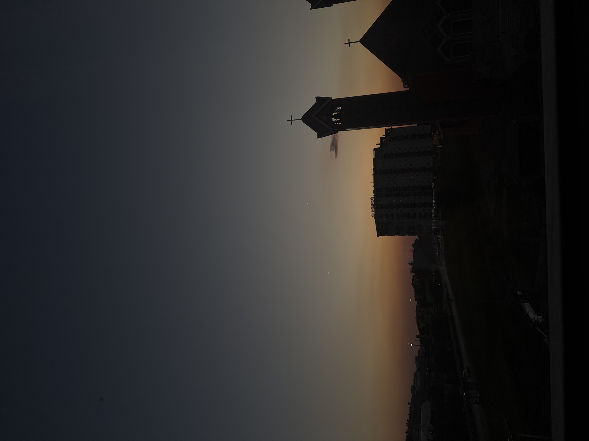 The image depicts a cityscape at dusk, possibly in Bar, Montenegro. A church silhouette with a visible cross stands against the fading light. Adjacent to the church, a tall residential building is discernible. The sky transitions from dark blue/grey to a warm orange/yellow near the horizon. Below the building, a street is visible with streetlights, and the landscape is generally obscured by darkness. The image orientation is sideways.