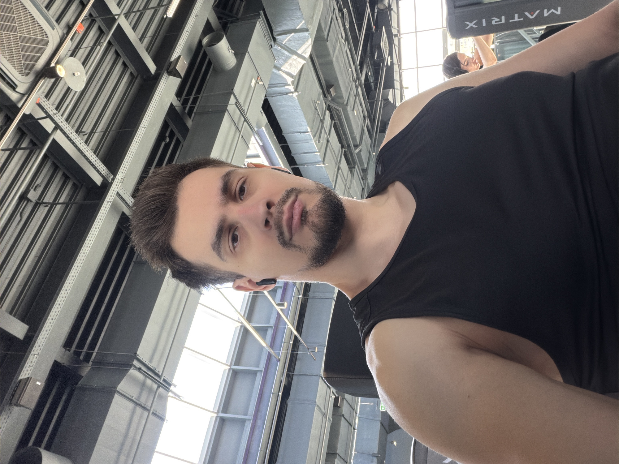 The image is a selfie taken at what appears to be a gym. The subject is a man with dark hair and a beard, wearing a black tank top. He has a small black earbud in one ear. The background shows gym equipment and a high ceiling with industrial architecture. The location is likely a fitness center in Krasnoyarsk, Russia, based on metadata associated with the image. A woman can be seen in the background near a Matrix treadmill.