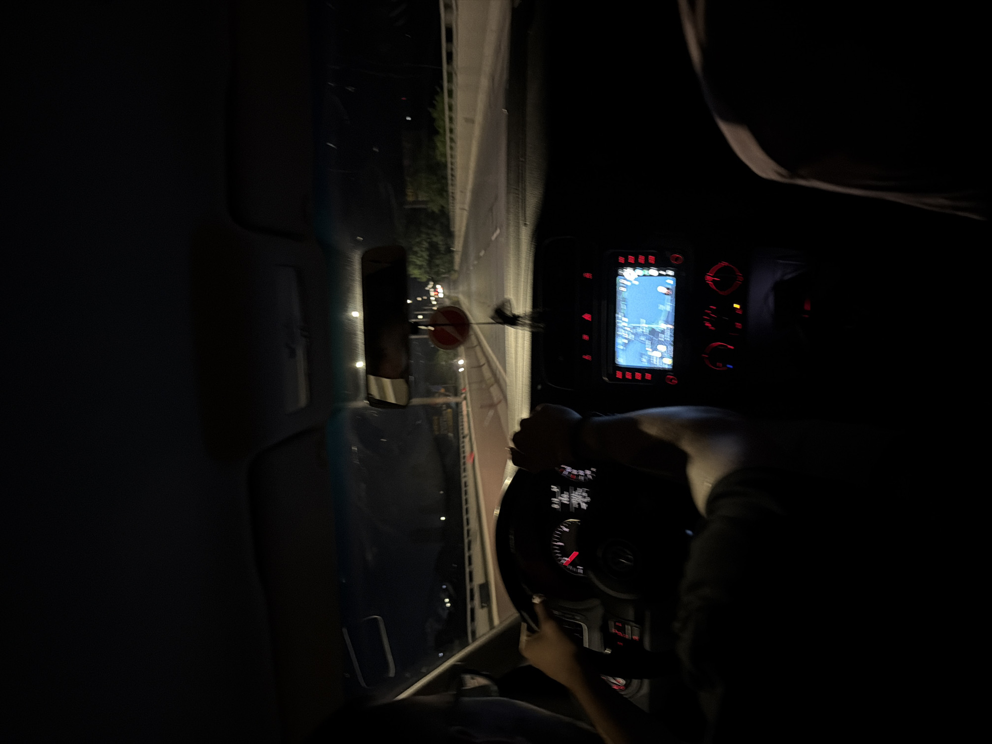 The image, rotated 90 degrees counter-clockwise, captures the interior of a vehicle at night, seemingly from a passenger's viewpoint. A person is driving, with their hands gripping the steering wheel, visible in the lower right of the frame. The driver's right arm and part of their torso are also discernible.
The car's dashboard is distinctly illuminated with red backlighting. A central screen displays a navigation map, rendered in blue and white tones, indicating a route or location. This screen is surrounded by control buttons and dials, also glowing red. The steering wheel features various controls and partially visible gauges (like a speedometer or tachometer) with red markings, obscured slightly by the driver's hands.
Through the windshield, a dark road unfolds, illuminated by distant streetlights and the faint glow of other vehicles' lights. It appears to be a multi-lane road or highway, possibly within an urban or suburban area like Burgas, Bulgaria. A circular red road sign with a horizontal white bar, typically signifying "no entry" or "do not enter," is visible on the right side of the road. Faintly lit trees or foliage line parts of the roadside. A rearview mirror is partially visible on the left, reflecting some ambient light. The scene is clearly set at night, indicated by the pervasive darkness and reliance on artificial light sources. No specific, legible text is clearly visible on the vehicle's displays or controls, only the general impression of numbers and symbols.