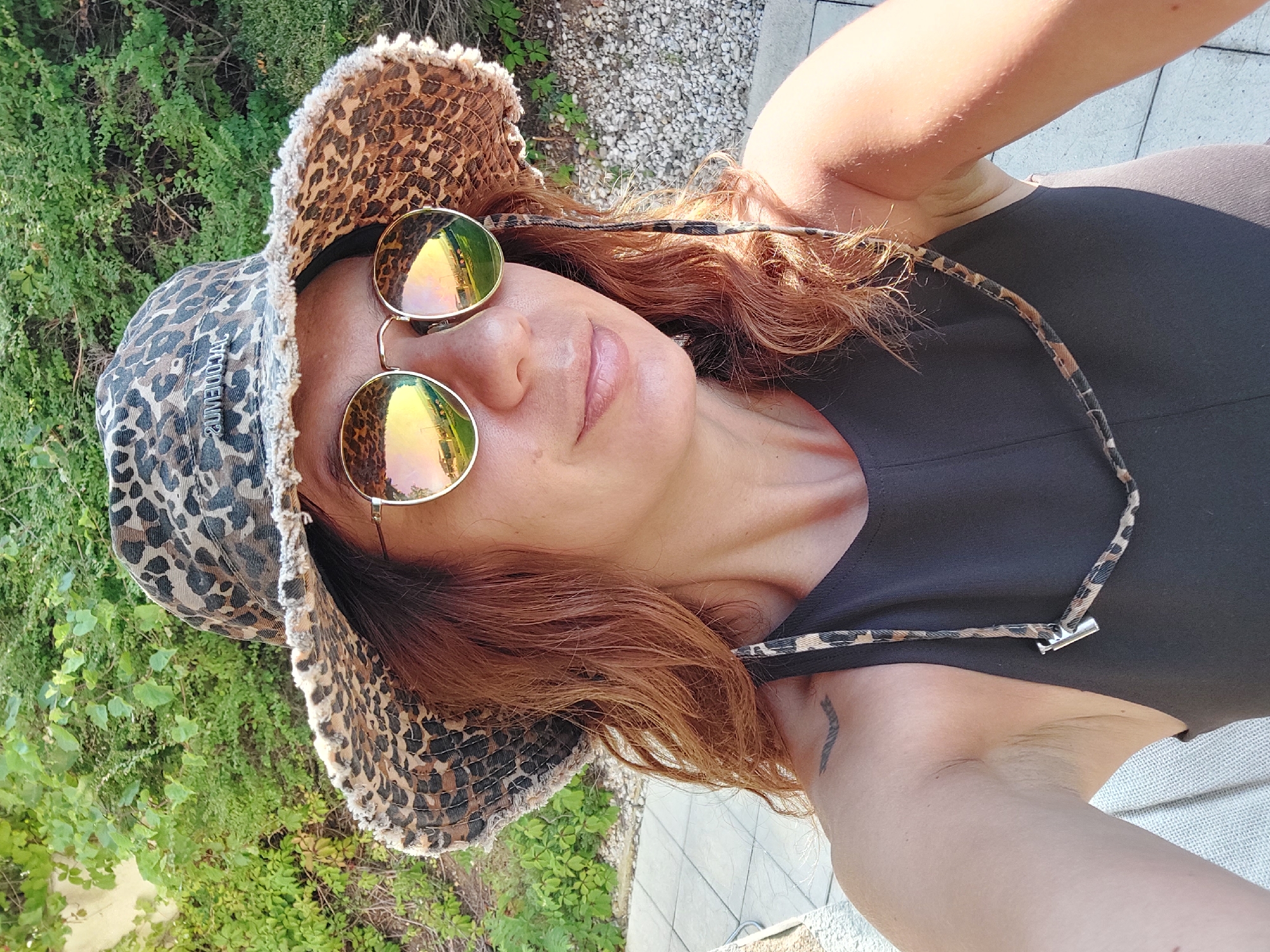 A woman is taking a selfie outdoors, wearing a leopard-print bucket hat and round, reflective sunglasses. Her reddish-brown hair is visible beneath the hat. She is wearing a dark sleeveless top, and a leopard-print strap with a silver clasp hangs from her neck. A small tattoo is visible on her left shoulder blade. The background consists of lush green foliage and some gravel. The paving stones in the lower left corner suggest an outdoor seating area, possibly a patio or terrace, within a location such as Sozopol, Bulgaria.