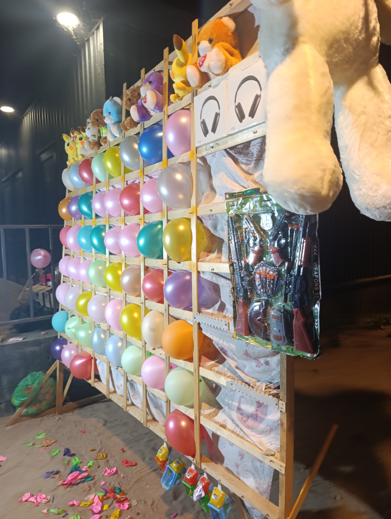 In Bosteri, Kyrgyzstan, a carnival game stall is set up, featuring a wooden rack filled with colorful balloons. Various plush toys, including teddy bears and Pikachu, are displayed above the balloons, seemingly as prizes. Below the balloons, a package of toy guns is hanging. The ground in front of the stall is littered with deflated balloons, suggesting the game has been played. The scene appears to be outdoors at dusk or night, with artificial lighting illuminating the stall.