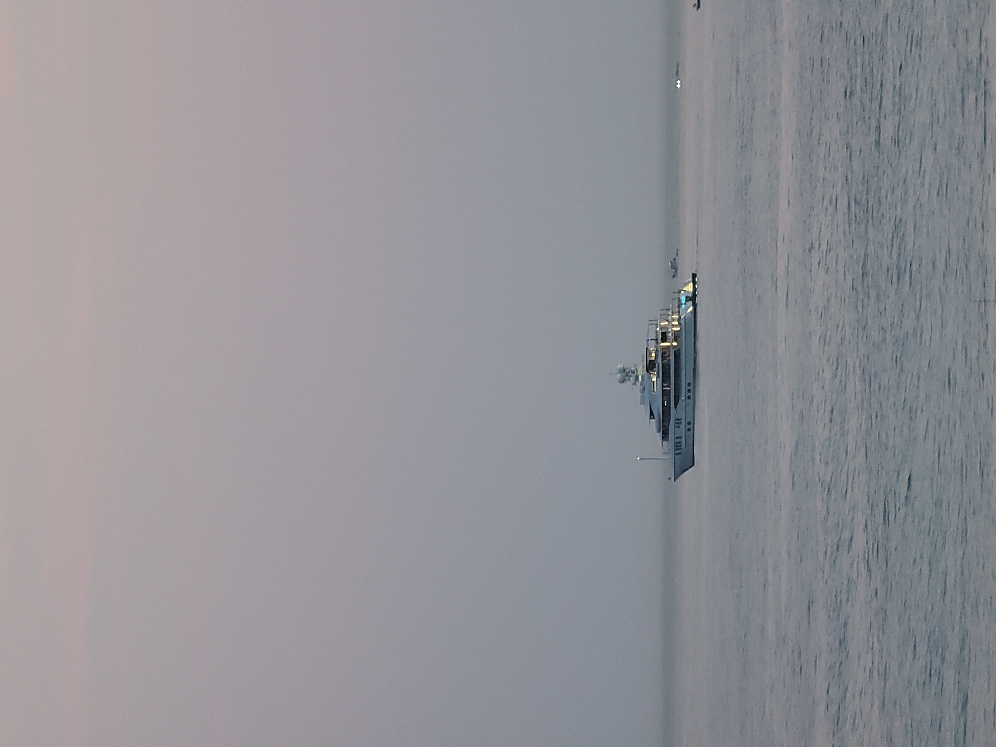 A large, multi-decked white yacht is sailing on a calm, grey sea under an overcast sky. The yacht has multiple lit windows, suggesting it is occupied. In the distance, to the right of the yacht, there are several smaller vessels. The scene appears to be set off the coast of Quarteira, Portugal, at dusk or early morning, indicated by the soft, diffused light. The water is a deep grey with subtle ripples, and the horizon is indistinct due to the hazy atmosphere.