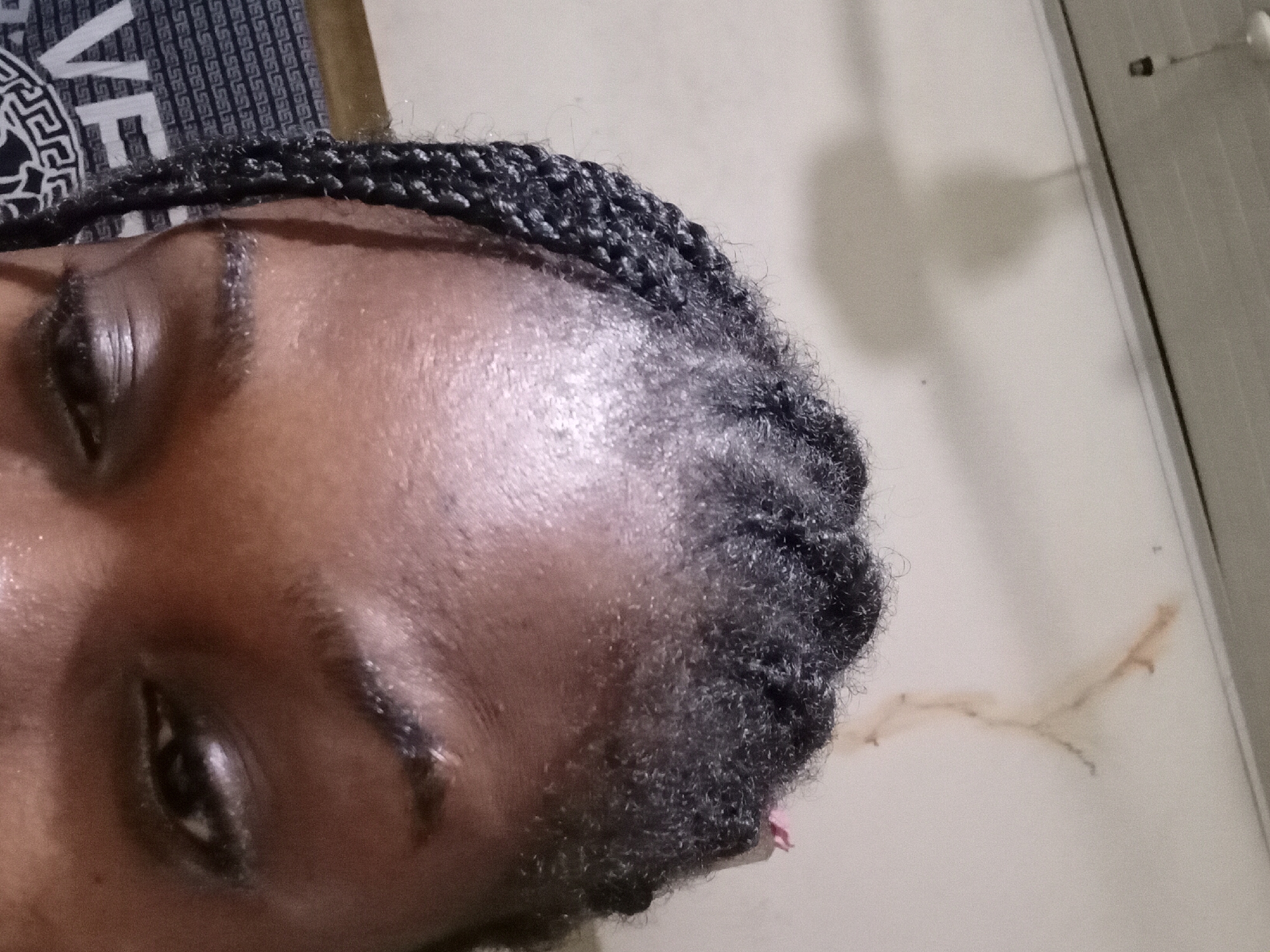 The image shows a close-up of a person's forehead, eyebrows, and eyes. The person has dark skin and their hair is styled in braids. The background appears to be an indoor setting with a wall that has some discoloration and a shadow that might be from a light fixture or a fan. There is also some text visible in the lower left corner of the image, which appears to be a logo or a design on clothing. The lighting is dim, and the focus is on the person's face. This image was captured in Ejule, Nigeria.