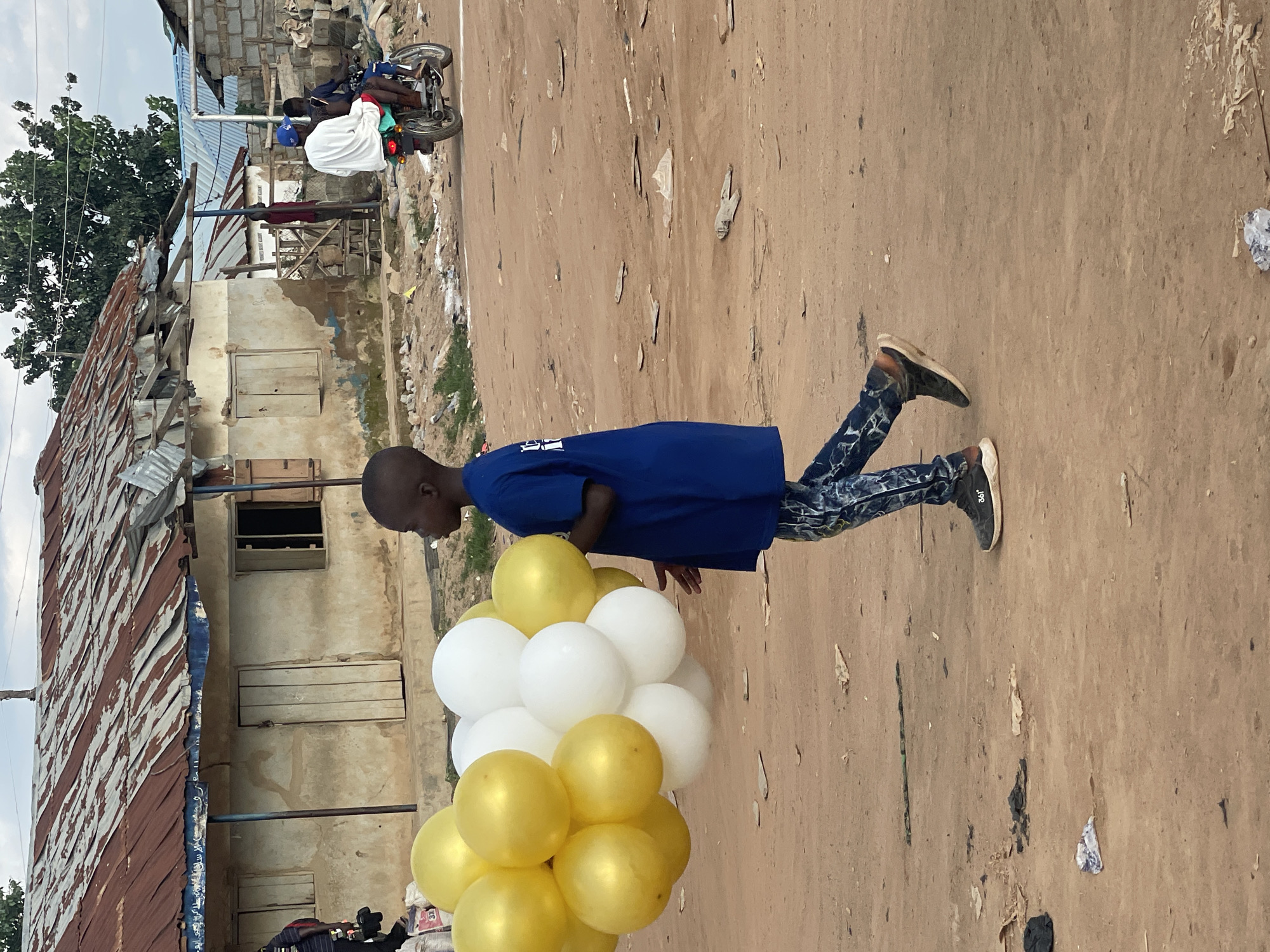 In Abuja, Nigeria, a young boy walks across a dirt road carrying a bouquet of yellow and white balloons. He is wearing a dark blue t-shirt and patterned pants. Behind him, a dilapidated building with a rusty corrugated metal roof stands under a cloudy sky. To the right of the building, two people are riding a motorcycle, and other buildings and people are visible in the background, suggesting a populated, possibly informal settlement. The ground is uneven and scattered with debris.