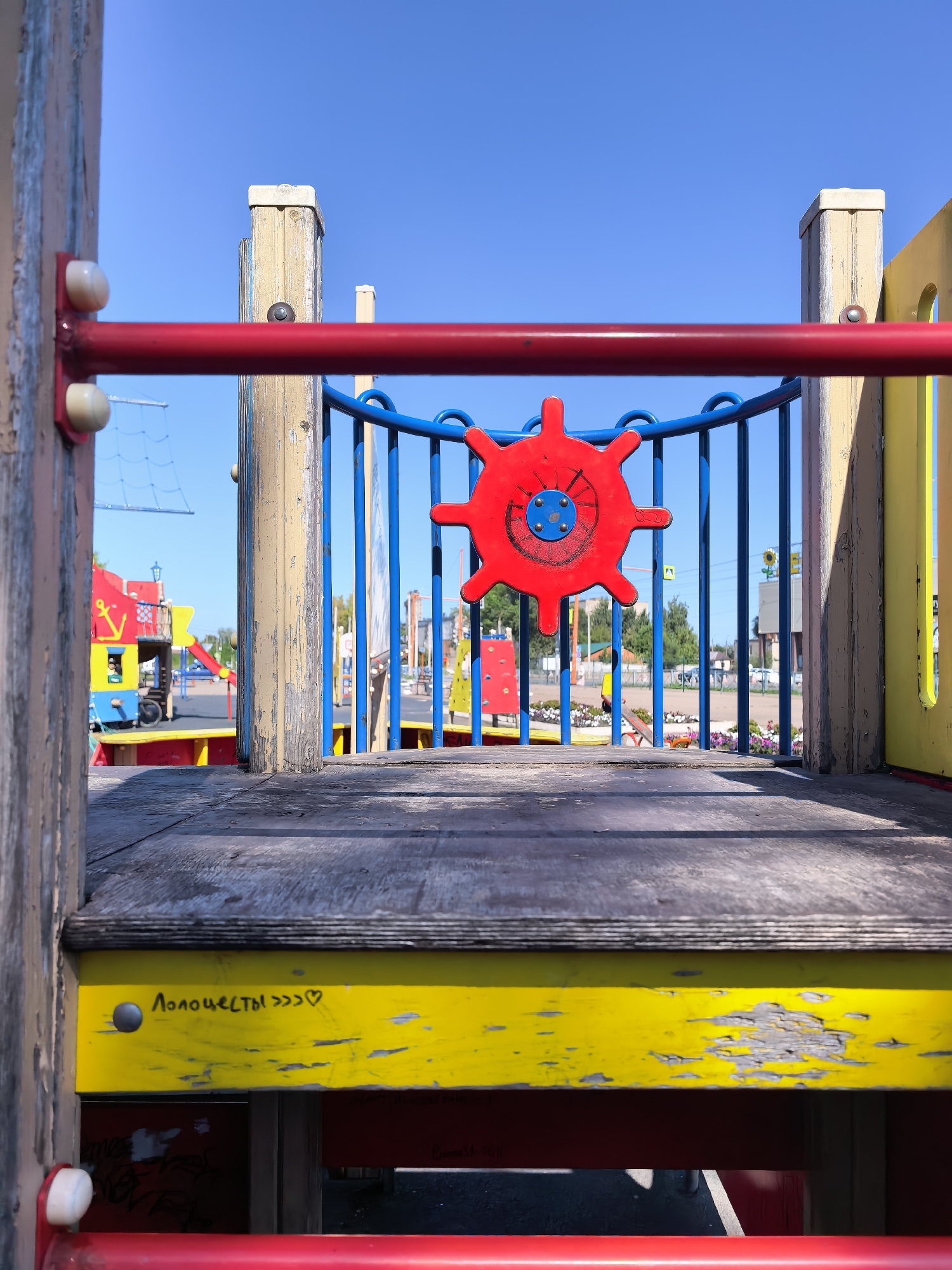 This is a close-up, eye-level, outdoor photograph of a brightly colored playground structure, under a clear blue sky. The foreground features weathered wooden planks and a red metal railing, with a yellow support beam visible below. A red ship's wheel, a common playground element, is centrally positioned in the mid-ground, attached to a blue metal railing. In the background, more playground equipment, including a red and yellow structure resembling a pirate ship with a slide, is visible. The scene is set in a public space, likely a park or schoolyard. The inscription "ЛЮБОЧЕСТЫ >>> ♡" is visible on the yellow beam. While the image is taken in Sterlitamak, Russia, there are no specific visual cues within the frame to pinpoint this exact location. No people are present in the immediate vicinity. The overall impression is one of a well-used, cheerful, outdoor play area on a sunny day.