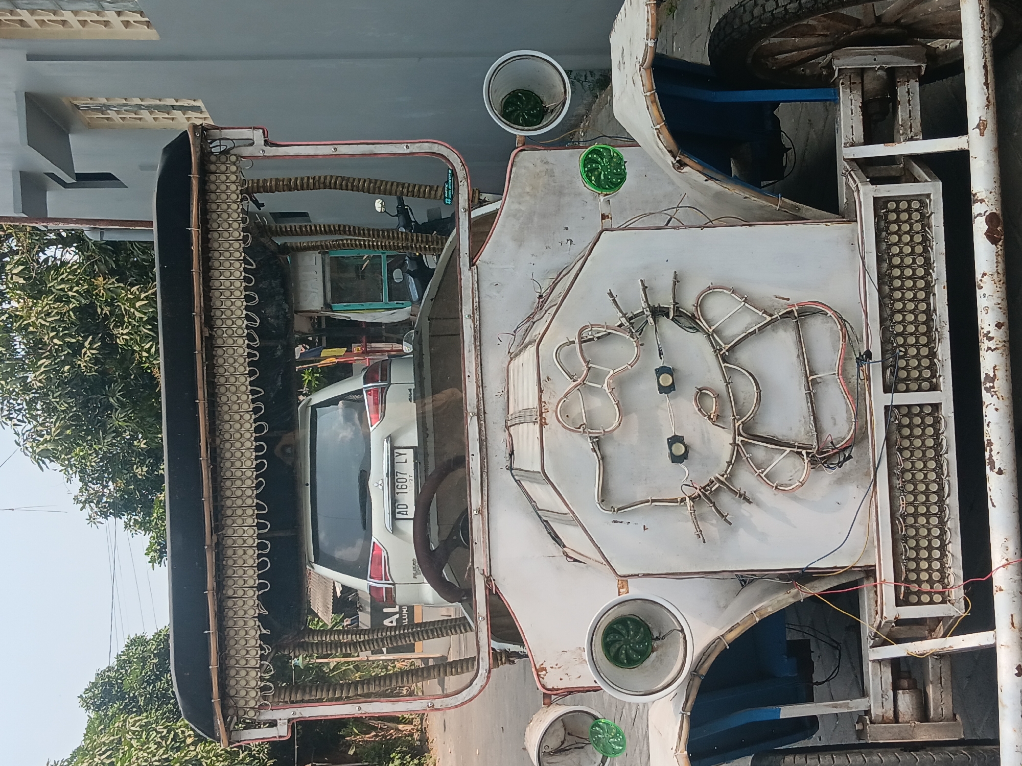 This image captures a street scene in Tlogo, Indonesia, focusing on a white, retro-style, open-top vehicle that appears to be a modified auto-rickshaw or a similar novelty vehicle. The vehicle's front is decorated with a large, illuminated outline of Hello Kitty. The vehicle has a rudimentary canopy supported by bamboo-like structures and adorned with rows of small, round lights. Inside the vehicle, the rear of a white Mitsubishi SUV with license plate "AD 1607 LY" is visible. The environment includes trees and a grey building with a textured facade. The scene appears to be a public street, with the vehicle likely parked or part of a parade or display.