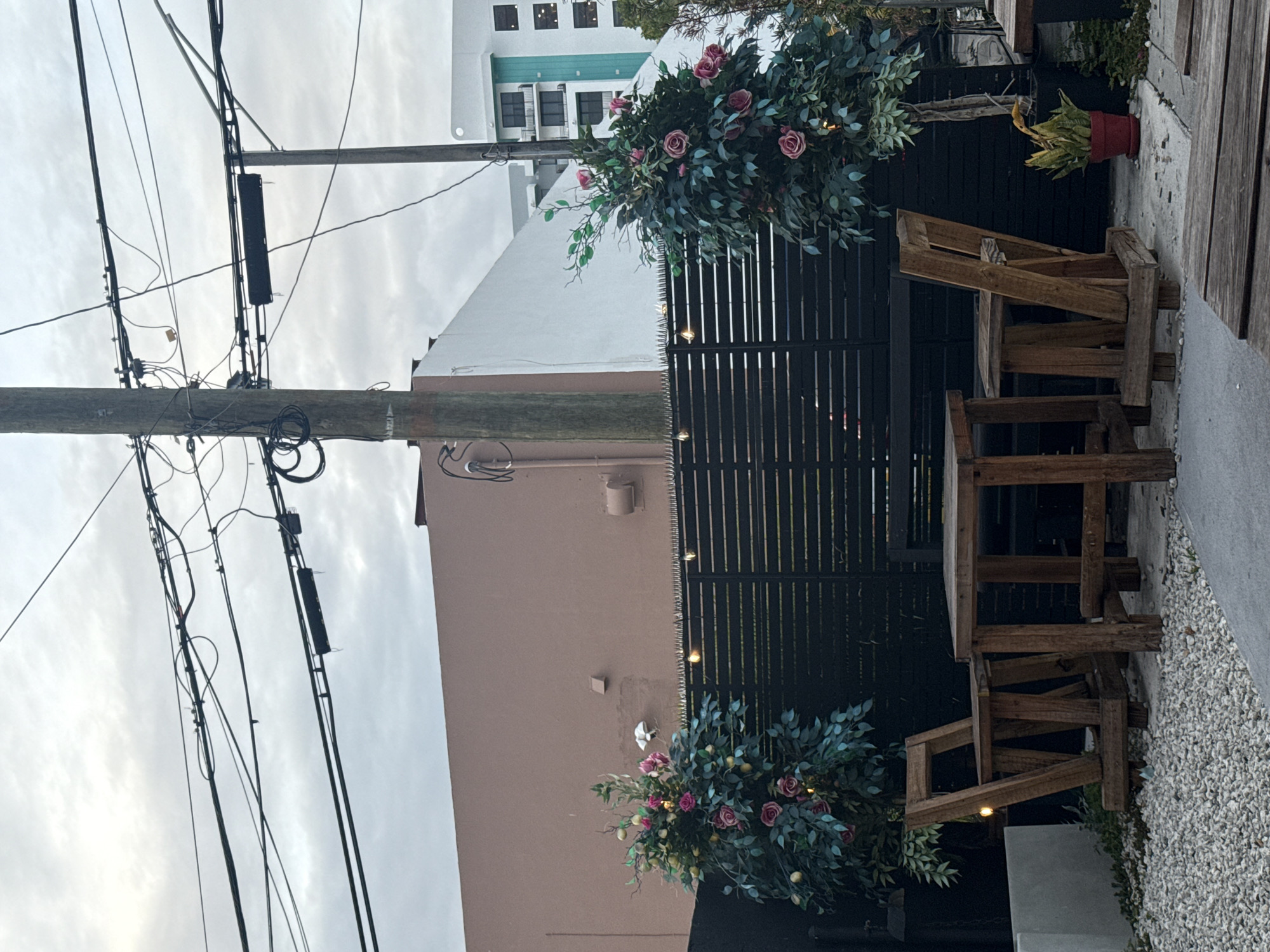 The image shows an outdoor dining area in Hollywood, United States of America, featuring a wooden table and chairs set against a black slatted fence decorated with floral arrangements. Above, utility poles and wires are visible against a cloudy sky. Buildings are visible in the background. The ground is covered in gravel.