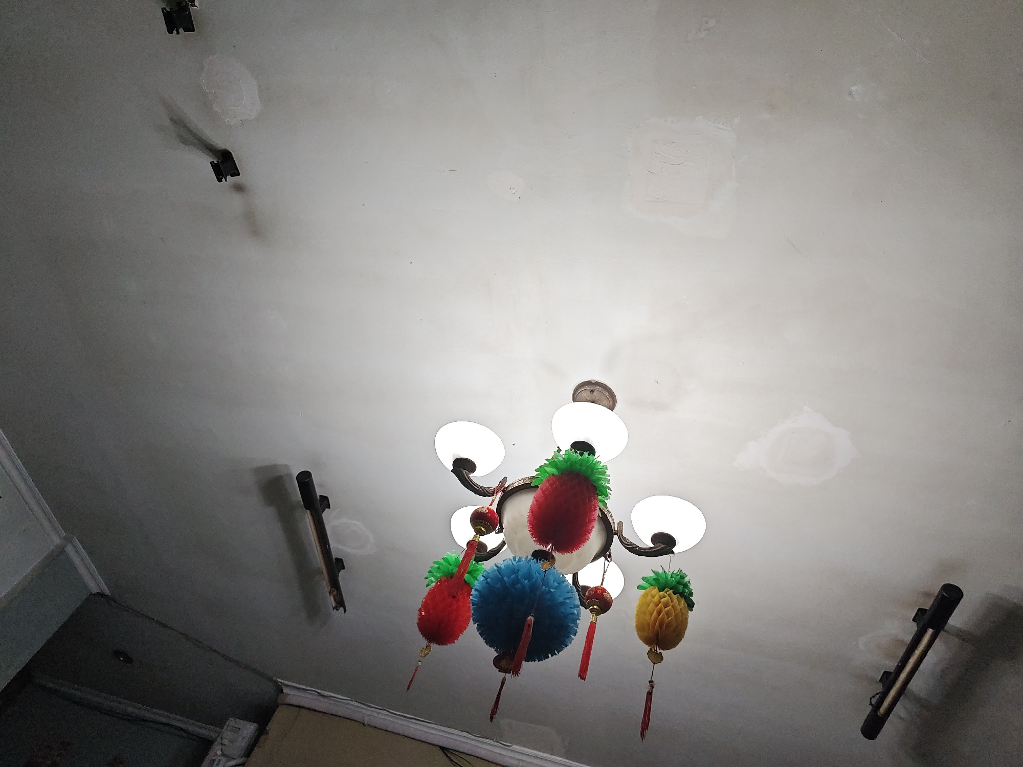 This is a low-angle, interior shot of a ceiling in North Jakarta, Indonesia. The ceiling is textured and shows signs of patching, with several circular and rectangular white patches visible. A chandelier with five white glass lamps hangs from the center. The chandelier has decorative elements shaped like pineapples in red, yellow, and blue, with green leafy tops and red tassels. Two dark, cylindrical objects with brackets are mounted on the ceiling on the left and right sides of the frame, casting shadows. The lighting appears to be artificial and uneven, with some areas of the ceiling brightly lit and others in shadow.