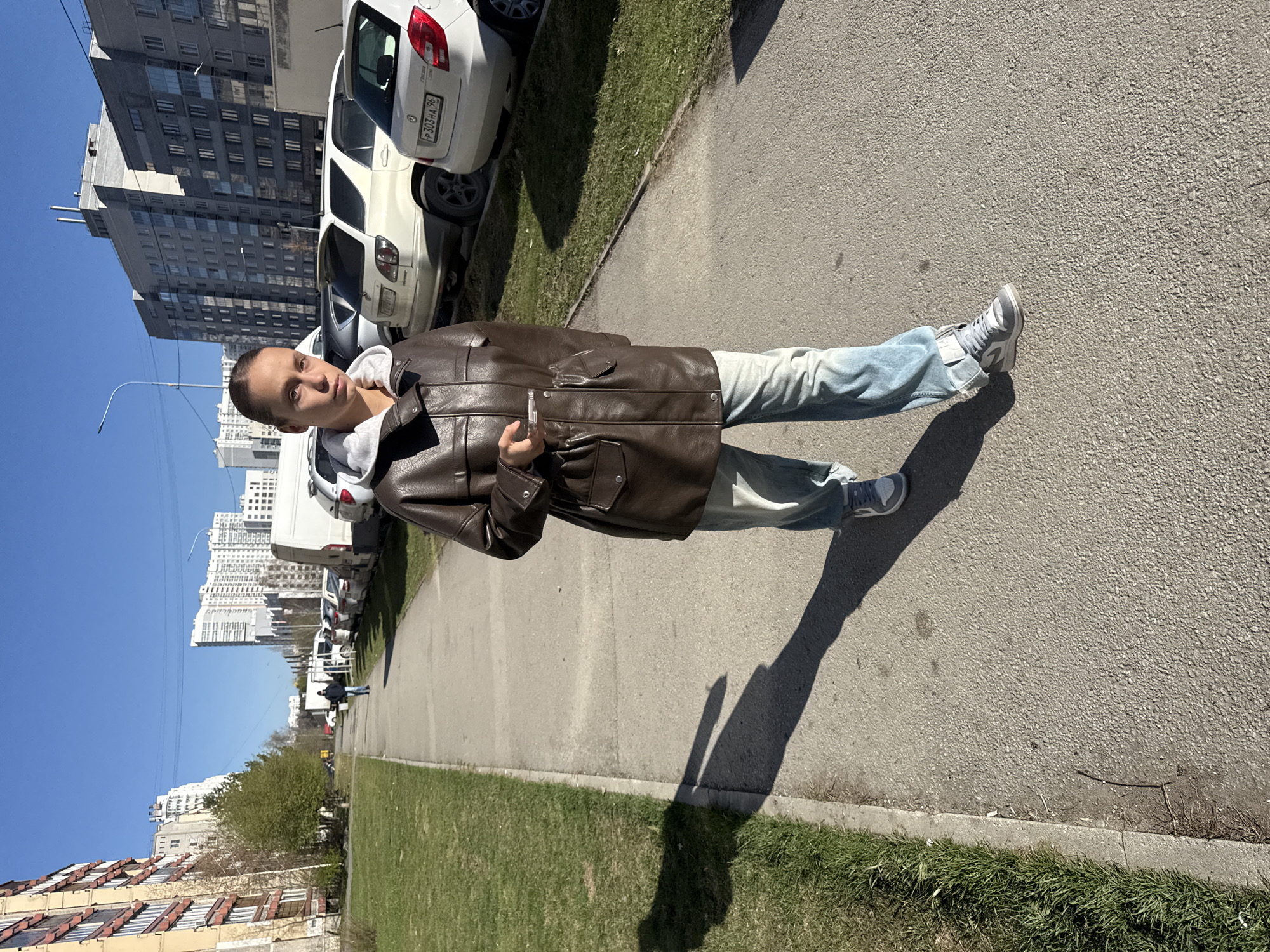 A young man stands on a sidewalk in what appears to be a residential area in Yekaterinburg, Russia. He is wearing a brown leather jacket, light blue jeans, and grey sneakers. He holds a small object in his hand, possibly a phone or electronic cigarette. The sidewalk borders a patch of green grass. Behind him, cars are parked along the street, and tall apartment buildings rise into a clear blue sky. The angle of the photograph is oblique, with the city skyline receding into the distance.