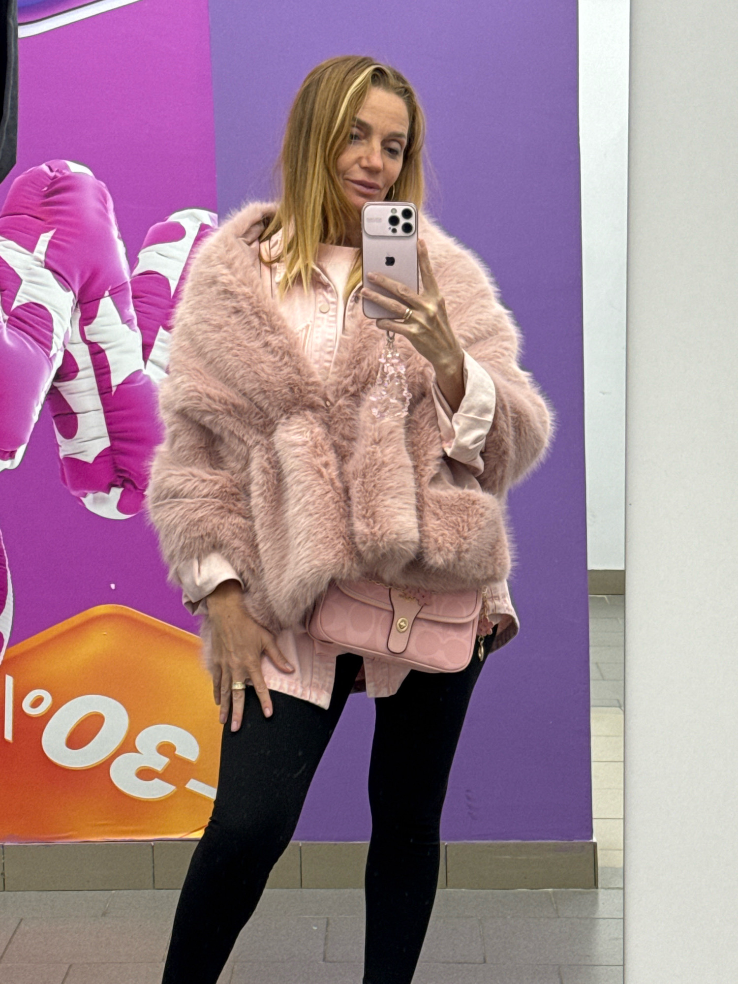 A woman is taking a mirror selfie in a brightly lit indoor space, possibly a mall or shopping center, in Захарьино, Russia. She is wearing a light pink faux fur coat over a pink button-down shirt, black leggings, and black shoes. A small pink crossbody bag with a subtle pattern is attached to her hip. She is holding a pink smartphone in her right hand, which is angled to capture the reflection. Her left hand rests on her thigh. In the background, a large, vibrant purple wall is visible, adorned with large, stylized pink and white balloon-like graphics. To the lower left, a portion of an orange and yellow sign with white text, including "30.⁰", is present. The flooring appears to be tiled in a light gray color.