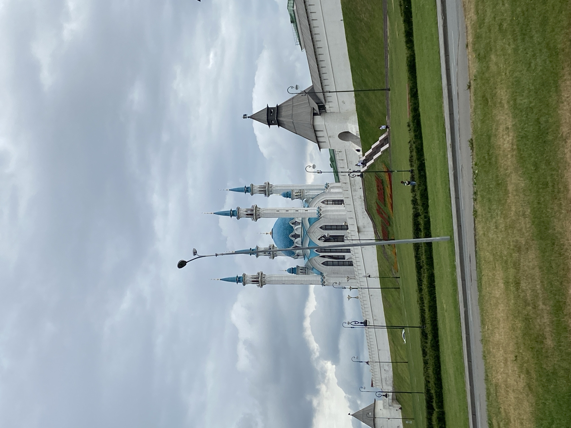 The image shows the Qolşärif Mosque in Kazan, Russia. The mosque is white with a blue dome and minarets. The sky is cloudy. There are lampposts along a lawn to the right of the mosque. A white wall with a tower can be seen in the background. Some people can be seen walking on steps near the tower. A road with green grass can be seen at the right edge of the image.