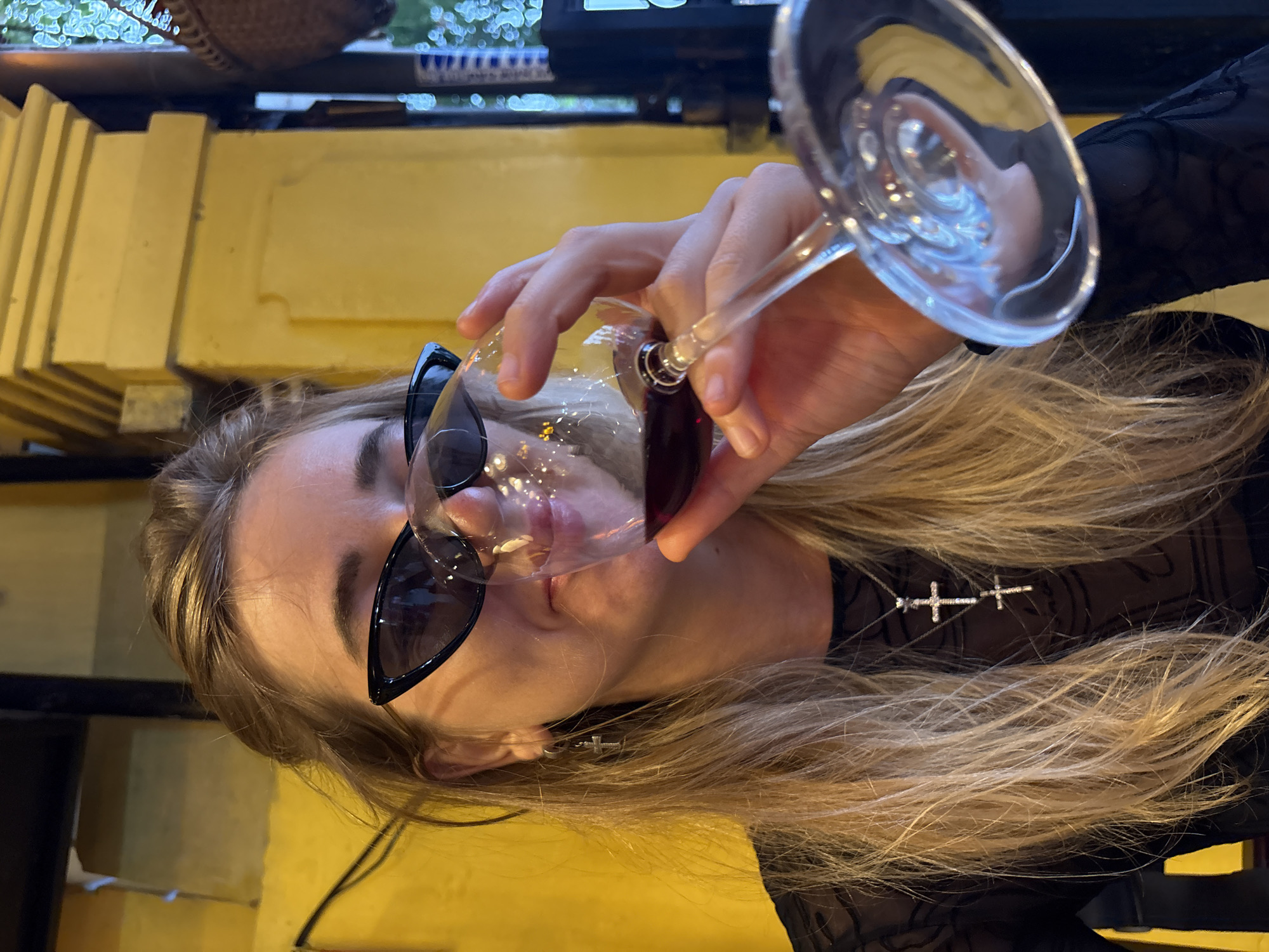 A person with long blonde hair and sunglasses is holding a wine glass filled with red wine, tilting the glass to drink. The person is wearing a black mesh top with a cross necklace. The background appears to be an outdoor setting with yellow walls and glimpses of greenery. The location is Hội An Ward, Vietnam.