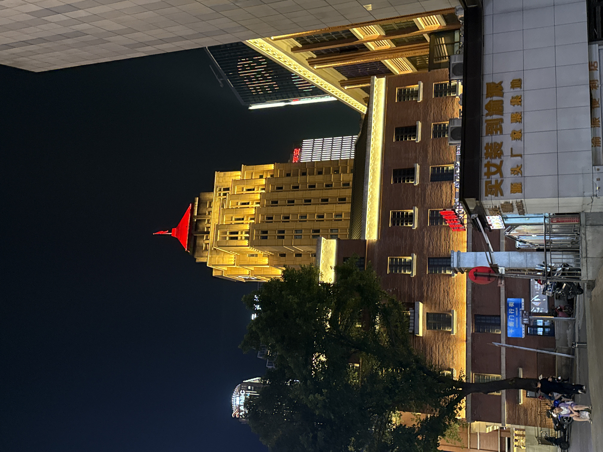 An exterior nighttime shot captures the illuminated architecture of Yuzhong District, China. The focal point is a tall, golden-hued building with a red-capped spire, its facade punctuated by rows of windows and ornate detailing, all bathed in warm, artificial light. Adjacent to it, a shorter, red-brick building features similar window arrangements and is also illuminated. In the foreground, a white structure with Chinese characters adorns its facade, suggesting a commercial establishment. A large, leafy tree stands to the left, partially obscuring the buildings. Several individuals are visible on the street in the lower left corner, some seated and others walking, with motorcycles parked nearby. The night sky is dark, providing a stark contrast to the illuminated structures.