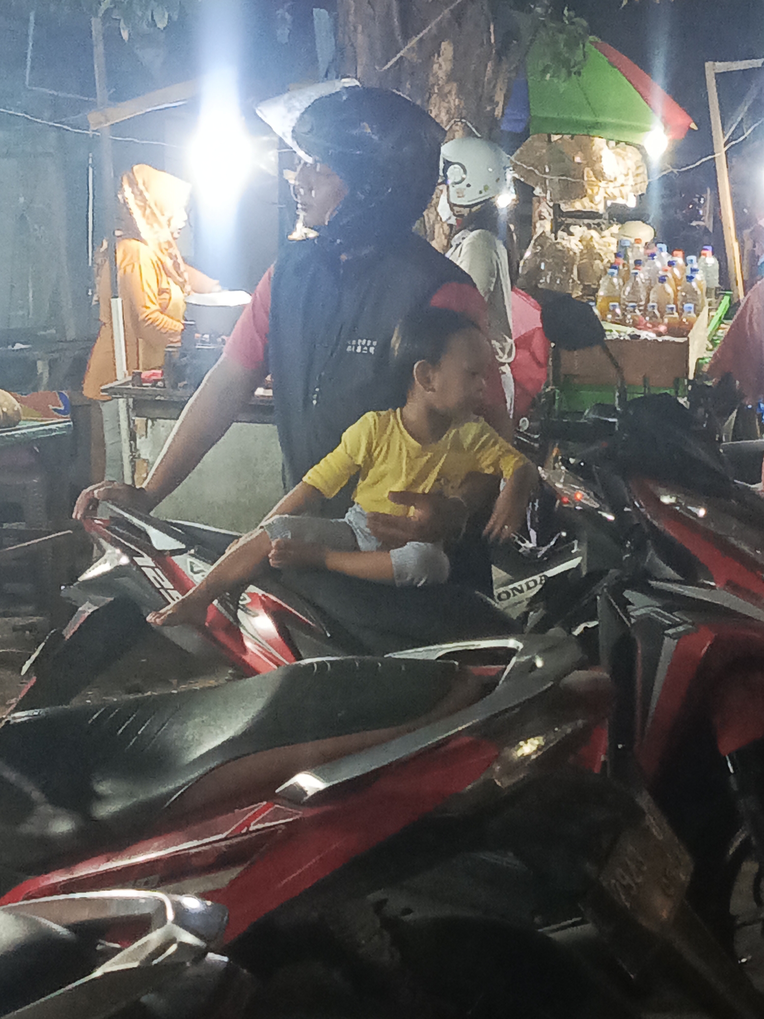 In Surabaya, Indonesia, at night, an adult wearing a black vest over a red shirt and a black helmet sits on a motorcycle. They are holding a young child, who is wearing a yellow t-shirt and grey shorts, on their lap. The child's legs are extended. In the background, other people are visible, including a woman wearing a headscarf and an orange top. There is a stall with bottles of liquid and food items. The scene appears to be outdoors, possibly on a street or market.
