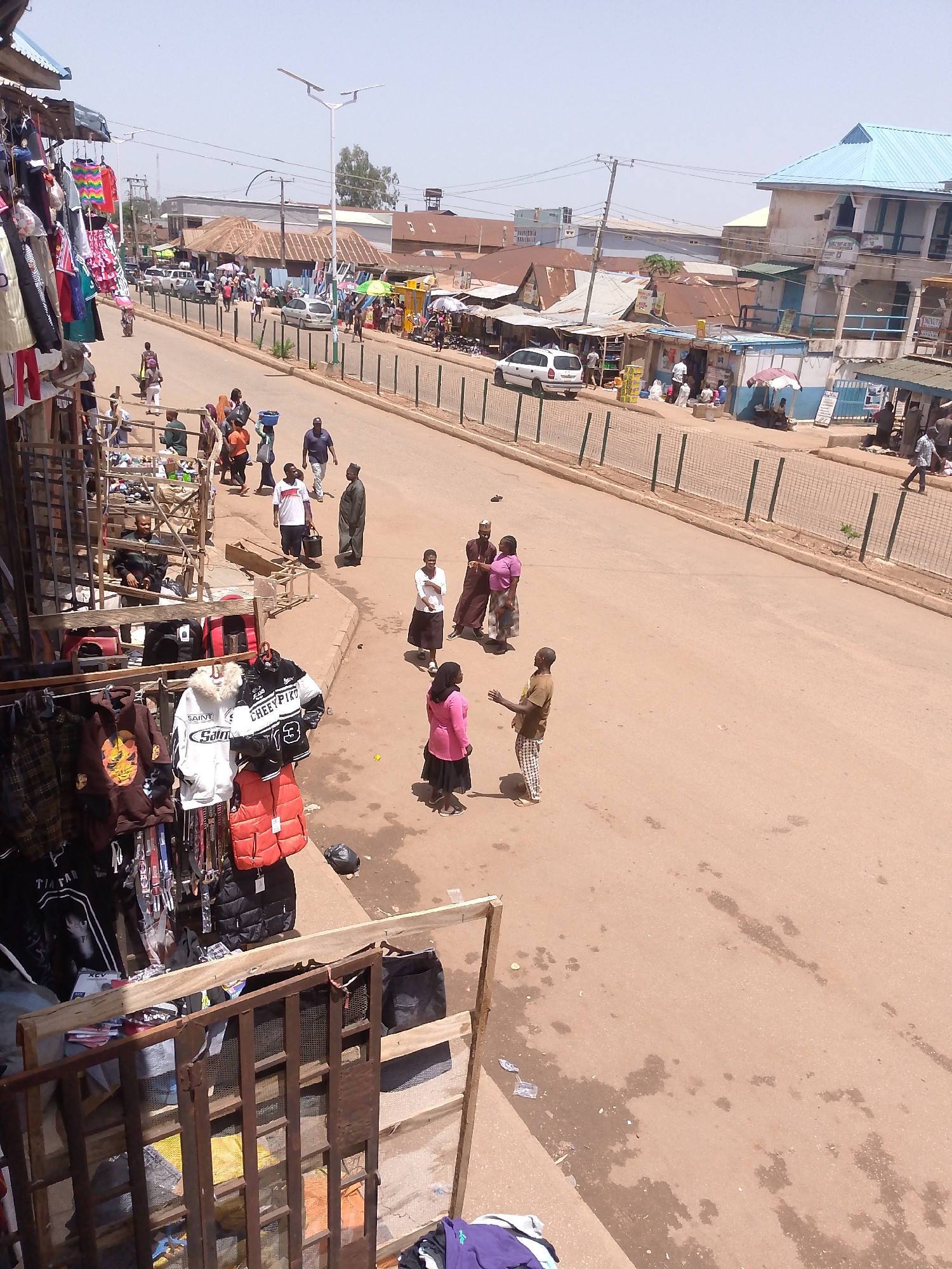 This photograph depicts a street scene in Bukuru, Nigeria. The road is asphalt, and several people are present. Some are walking, others are standing and talking in small groups. On the left side of the road, there is a shop with clothing and other goods displayed on a rack. Across the road, there is a fence and a line of buildings with varying roof types. Vehicles are parked along the street. Overhead, there are utility poles and wires. The sky appears to be clear.