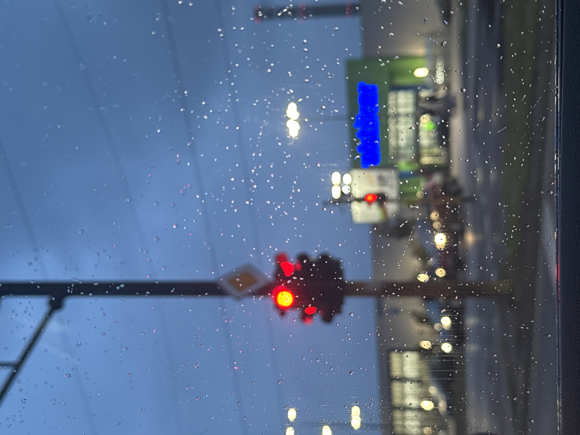 The image shows a view through a rain-streaked window, likely a car windshield. Outside, a blurred street scene is visible at night. A traffic light is red. Further away are streetlights and a building with a bright green and blue sign. Cars are lined up on the right, with their headlights illuminated. The image might have been taken from within a stationary vehicle. Location might be Sofia, Bulgaria but can not be confirmed.