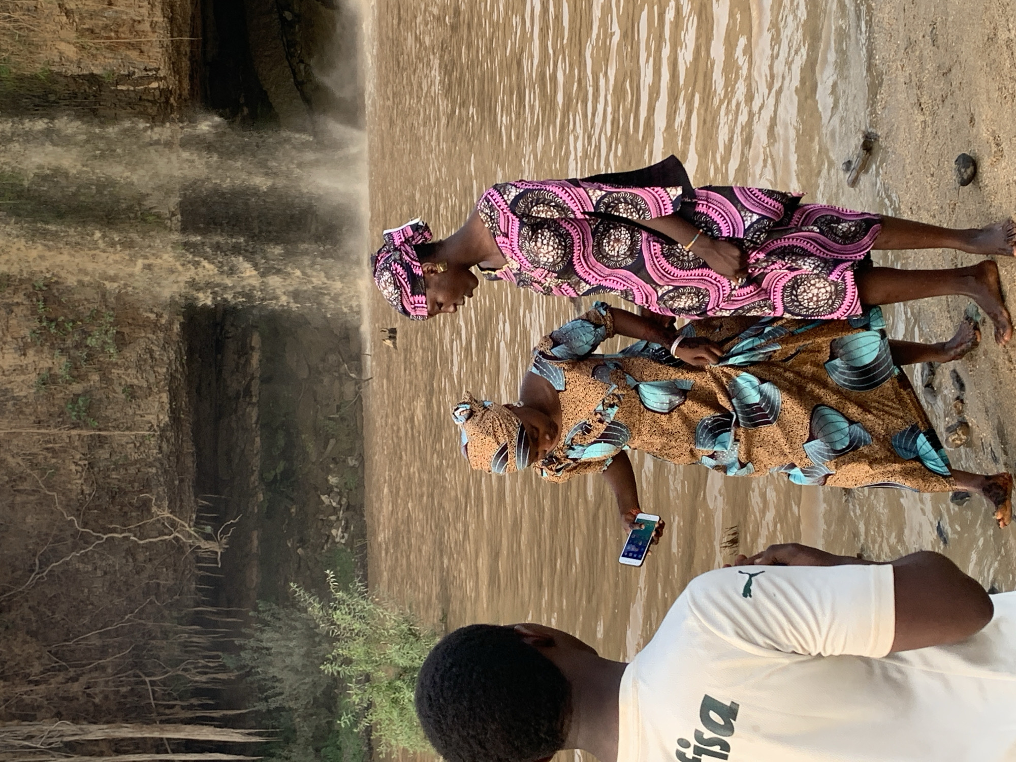 Two women and a man stand near Gurara Falls in Kukum, Nigeria. The falls are on the left. The women are wearing patterned dresses and head coverings, with the woman in the front holding a smartphone. The man is wearing a white shirt with a logo on the front. The ground appears to be sandy and wet.