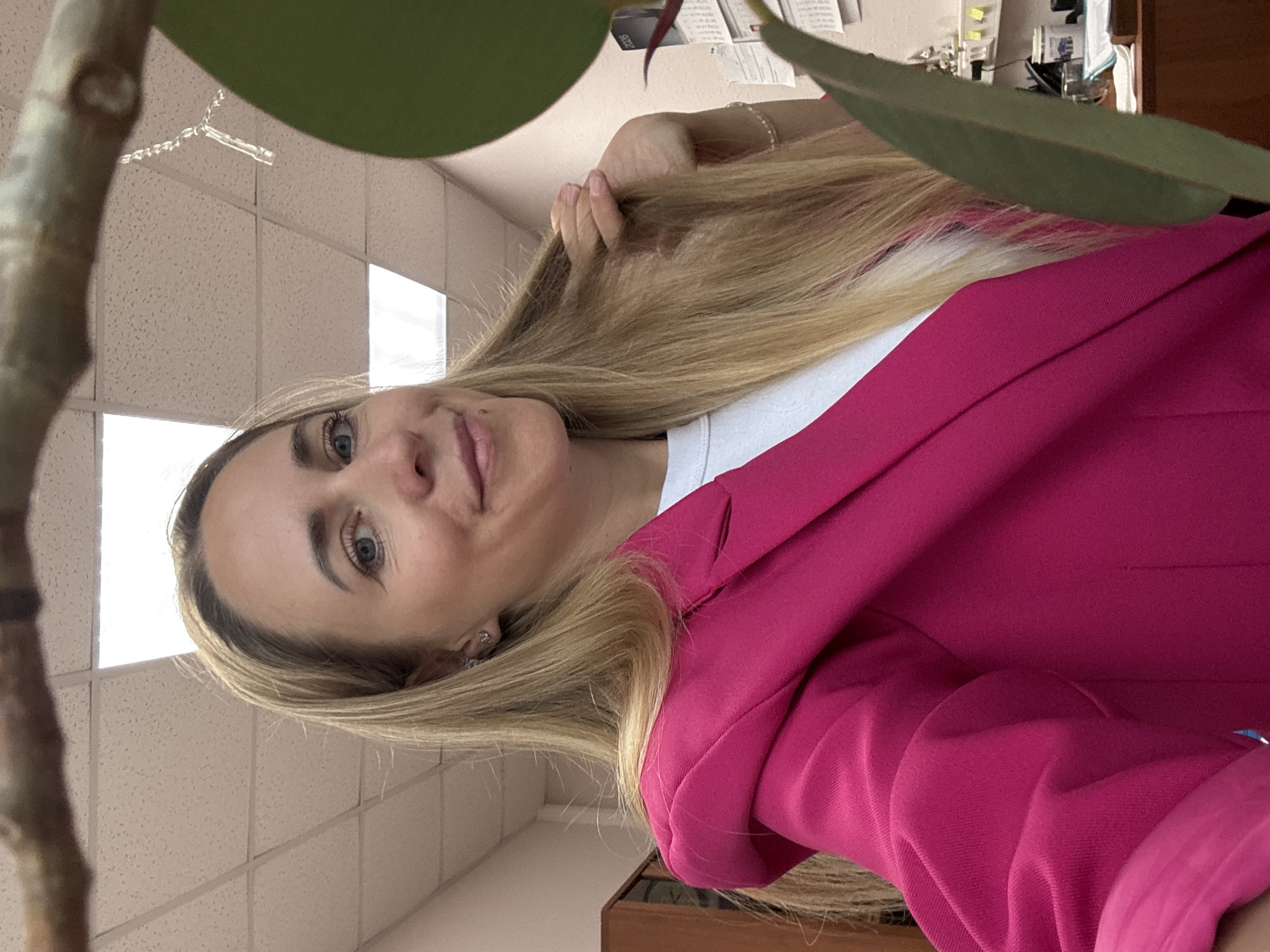 A woman with blonde hair and blue eyes is taking a selfie in an office environment. She is wearing a bright pink blazer over a white shirt. Her left hand is resting on her hair, and she is smiling. In the foreground, a large green leaf from a plant partially obscures her right side. In the background, ceiling lights are visible, along with some office furniture and a filing cabinet. A branch with some delicate, string-like objects hangs from the top of the frame. The scene appears to be indoors, likely in a workplace. The image does not provide any specific details to indicate its location in Chaikovsky, Russia.