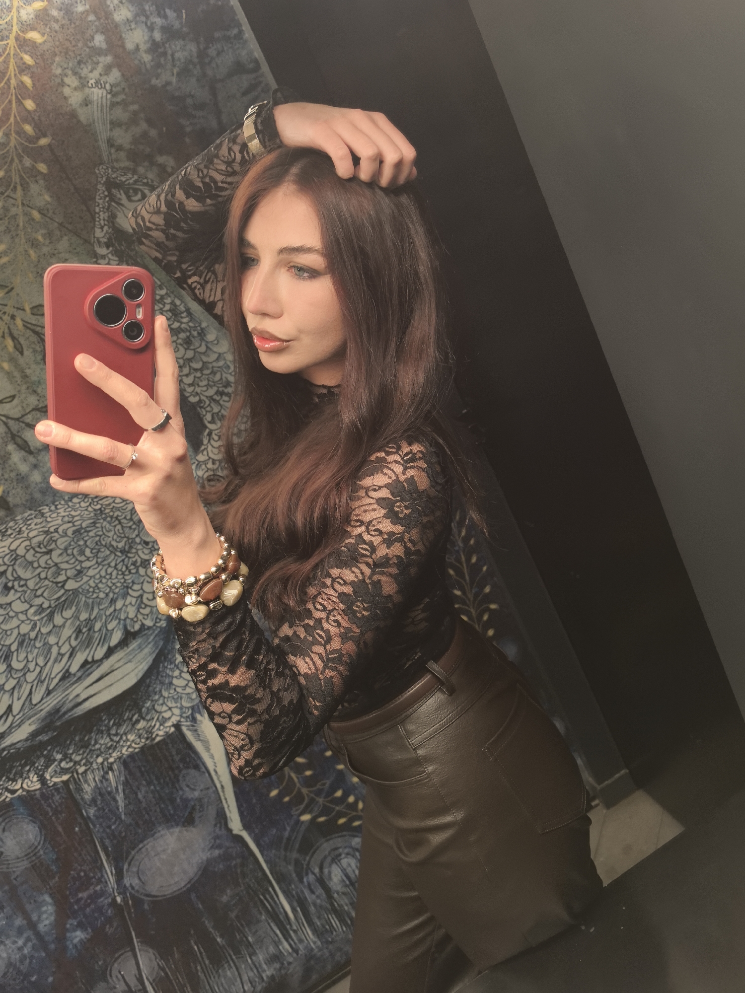 A person takes a mirror selfie in what appears to be a lavatory. The person has long, wavy, auburn hair, fair skin, and blue eyes. They are wearing a black lace top, brown leather pants, and bracelets on their left wrist. They are holding a red smartphone in their left hand, with two rings on their fingers. The backdrop consists of a wall with a blue and white pattern resembling a bird or a floral motif on the left and a plain dark gray wall on the right. The location details, including the city of Stara Zagora, Bulgaria, are not explicitly evident in the visual content.