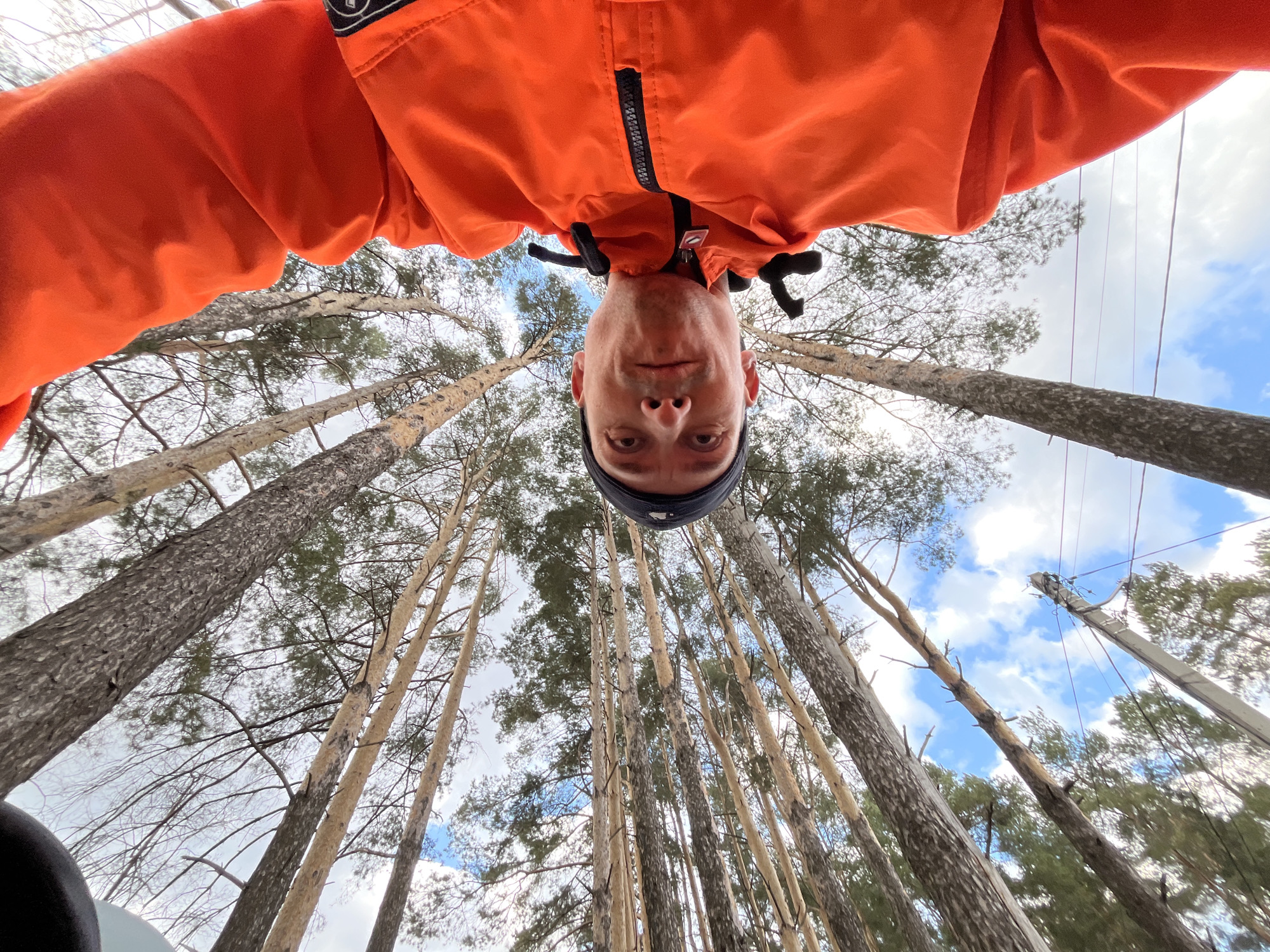 An unusual, low-angle selfie captures a man appearing upside down against a backdrop of tall trees in Chaikovsky, Russia. The man's face and upper torso are visible from below, as if he is looking down from above. He wears a vibrant orange jacket with a black zipper and a dark beanie, with a small, illegible logo or patch visible on his left shoulder. His expression is neutral as he looks directly into the camera.
The background is dominated by numerous tall, slender trees, resembling pines, which stretch upwards towards a partly cloudy sky. The perspective is taken from directly below, emphasizing the height of the trees and creating a sense of immersion within the forest. Patches of blue sky are visible between the tree branches and clouds. On the right side of the frame, a utility pole with power lines extends upwards, contrasting with the natural environment. The scene is brightly lit, indicating it is daytime. The image presents a creative and playful self-portrait taken in a wooded outdoor setting.