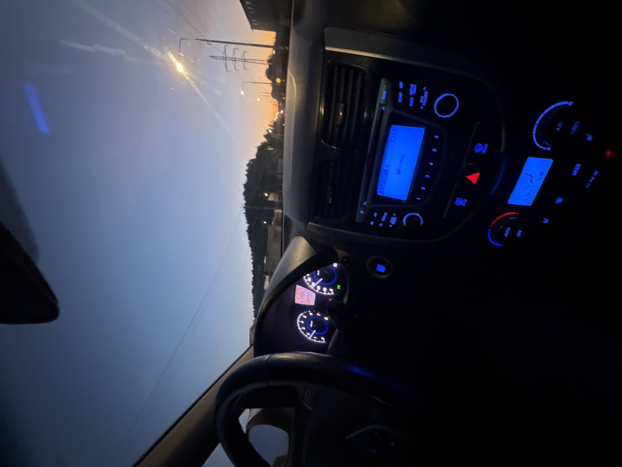 This media file presents a view from inside a car during twilight or late evening, rotated 90 degrees clockwise. The image captures the illuminated dashboard and the fading light through the windshield.
Inside the car, the infotainment system is prominently lit with blue backlighting, displaying "23:00" as the time and "MP3 Play" as the current mode. Other visible buttons and labels on the console include "SCAN", "INFO", "SETUP", "FOLDER", "MODE", "A/C", "AUTO", "OFF", "CD/AUX", and numerical buttons "1" through "6". A red triangular hazard light button is visible, along with a climate control display showing "22.0C" for the temperature and a "FRONT" vent button. Part of the instrument cluster is visible behind the steering wheel, also illuminated with white and blue lights. The steering wheel itself is partially visible in the foreground.
Outside the car, the sky presents a gradient of deep blue transitioning to an orange-yellow glow near the horizon, characteristic of sunset or deep twilight. A bright streetlight in the distance emits a starburst glare, and silhouettes of power lines, trees, and distant structures are visible against the colorful sky. The clear sky suggests calm weather. The car appears to be driving or parked on a road in Chaikovsky, Russia, with the time display (23:00) confirming the late hour.