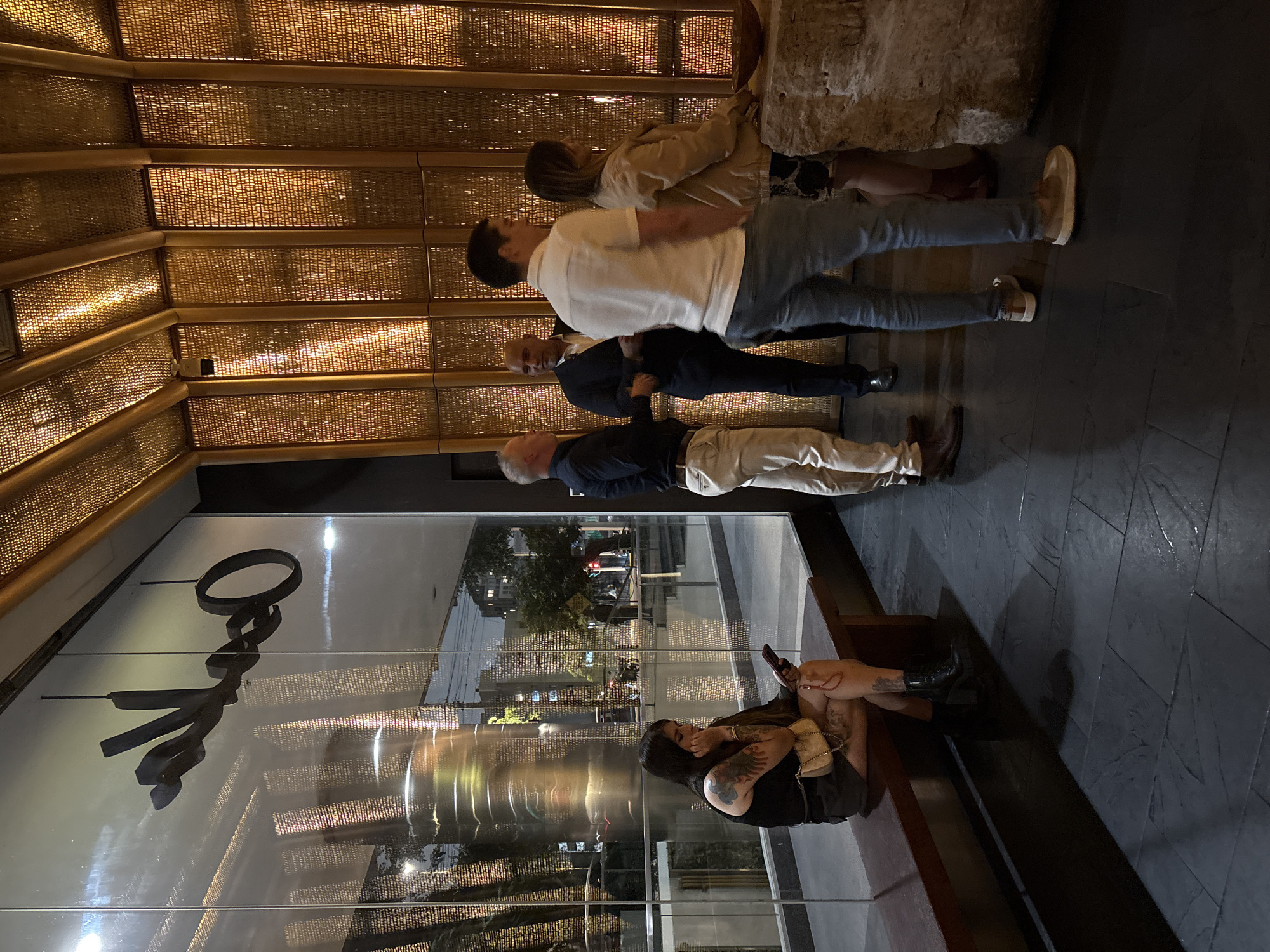 In the city of San Isidro, Peru, outside a establishment with a reflective glass facade featuring the stylized word "Ophelia" in cursive, several individuals are visible. To the left, a woman with tattoos on her arms and legs is seated on a dark brown wooden bench, looking down at her phone. She is wearing a black tank top, a dark skirt, and black boots. A small, light-colored crossbody bag is on her lap.
To the right, four individuals are standing near a textured, golden wall and a large, light-colored stone structure. Two men in the foreground are engaged in conversation. One man, facing away from the camera, wears a white short-sleeved shirt and light blue jeans. Another man, with dark hair and a dark suit, is speaking to a man in a dark blue shirt and light-colored pants. A woman in a tan jacket stands to the right of the man in the white shirt. The floor is a dark, tiled surface with visible reflections. The lighting suggests it is evening or nighttime, with artificial light illuminating the interior of the establishment and casting shadows.