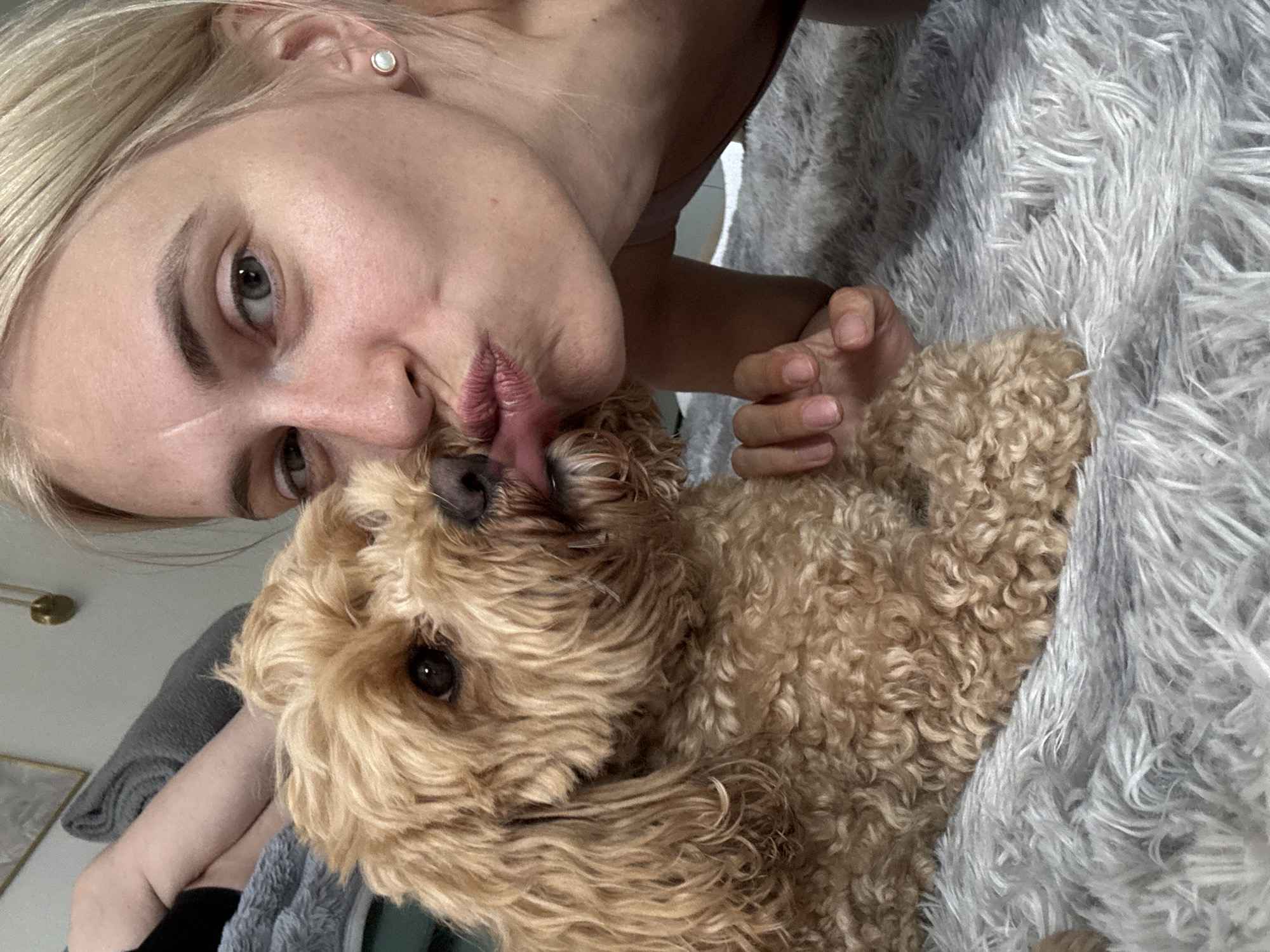 The image shows a close-up, slightly angled shot of a woman and a dog. The woman has blonde hair and is wearing a light-colored top. She is leaning in towards the dog, with her lips pursed as if to kiss it. Her left eye is visible, along with part of her nose and forehead. She has a scar on her forehead. The dog is a light brown color with curly fur, and its face is positioned near the woman's. The dog's right eye is visible, and its tongue is slightly out, touching the woman's mouth.
The background appears to be a soft, fluffy gray blanket or bedspread. Some of the woman's arm and hand are visible holding the dog. The overall lighting suggests it might be daytime, possibly indoors, as there are no strong outdoor light cues. The emotions conveyed are affection and playfulness between the woman and her pet. The setting appears to be a domestic environment, likely a bedroom or living area, given the soft furnishings. There is no visible text in the image.