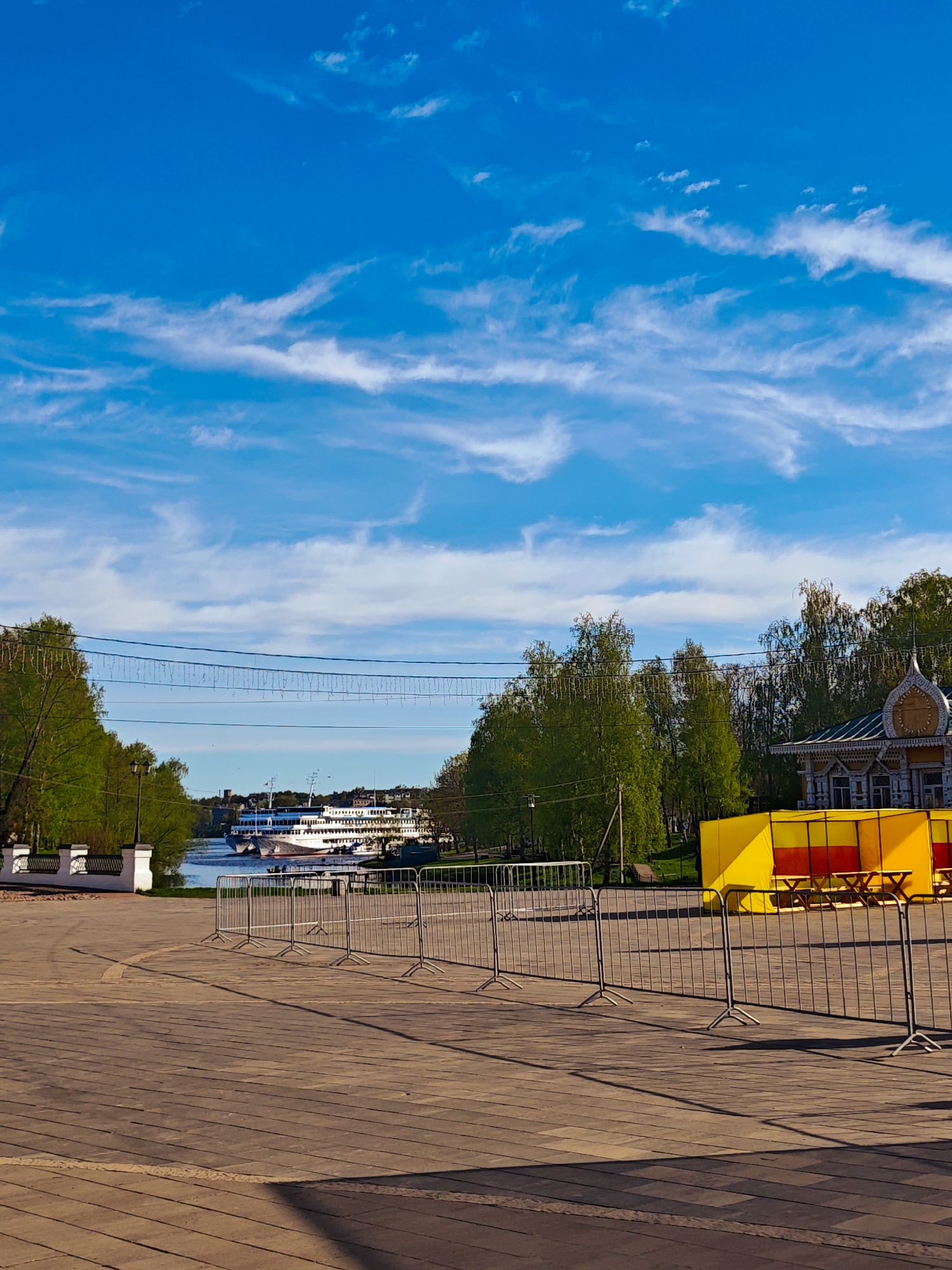 The image depicts a town square in Chelganovo, Russia, on a sunny day with a blue sky dotted with wispy white clouds. The square is paved with wooden planks. A metal barricade runs across the foreground, separating the viewer from the rest of the scene. Behind the barricade, there is a grassy area with trees and a decorative building with intricate wooden carvings. In the background, a large passenger ship is docked on a river or lake. A yellow and red structure is positioned to the right of the image, seemingly enclosing picnic tables. The scene suggests a public space prepared for an event, with the barricade potentially serving as a crowd control measure.