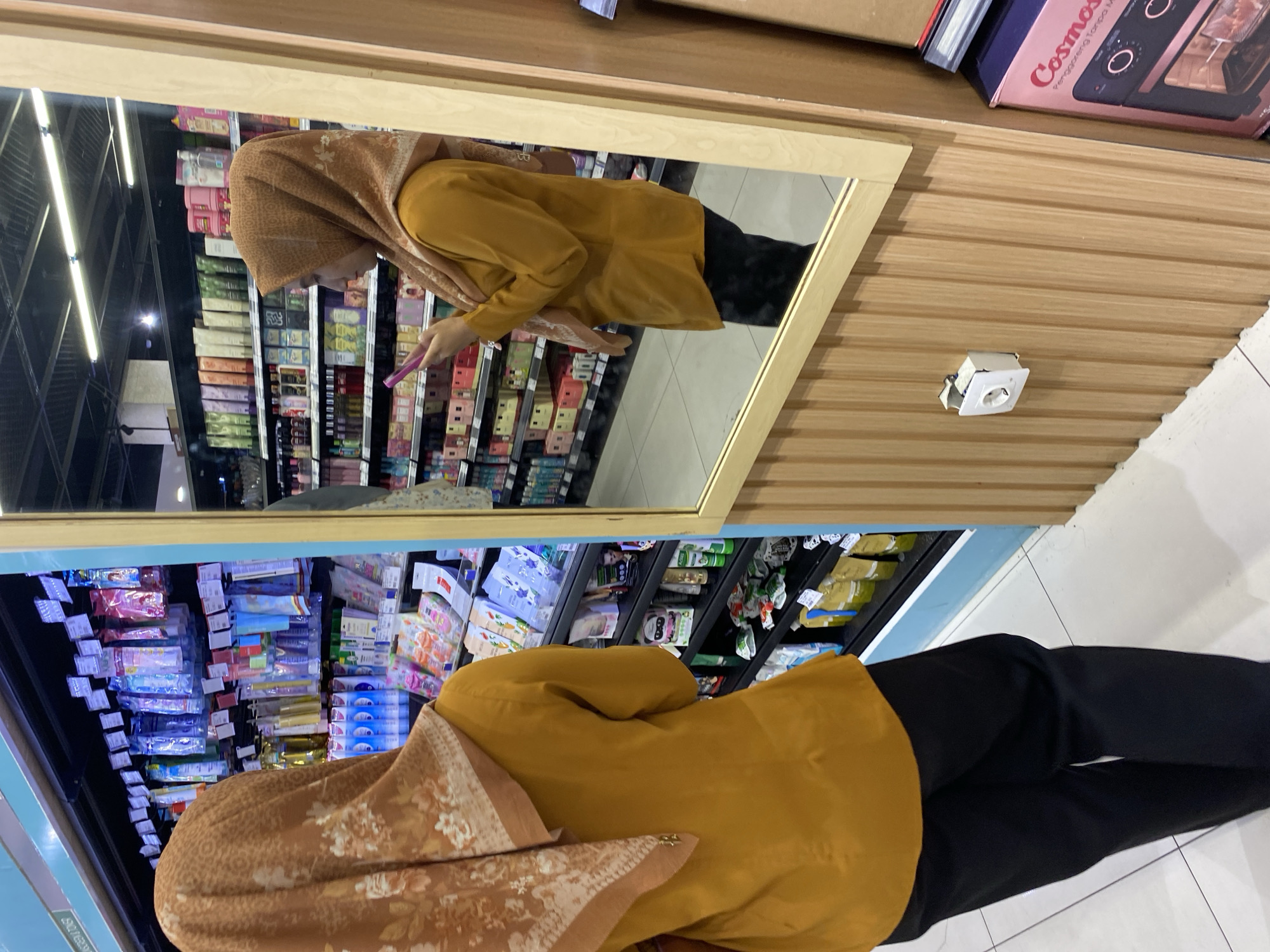 A woman wearing a mustard-colored tunic and black pants, with a patterned hijab, is standing in a supermarket aisle and looking at her phone, reflected in a mirror. The aisle is stocked with various personal care products, including tubes of cream and packets of facial masks. The mirror is framed with light-colored wood paneling. To the right of the mirror is a large pink box containing what appears to be a kitchen appliance. A white electrical outlet is visible on the wood paneling below the mirror. The floor is tiled. The image captures a moment of everyday life in a retail setting.