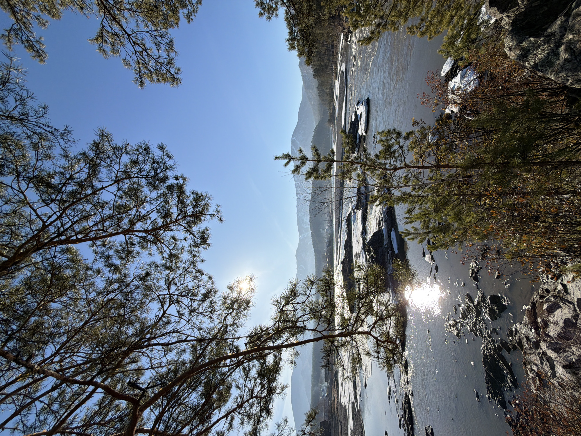 The image captures a landscape in the city of Manzherok, Russia, featuring a partially frozen river with mountain ranges in the background. The foreground includes trees with bare branches. The sky is blue, and the sun is visible, reflecting off the water. Rocks and patches of ice are present along the river's edge.