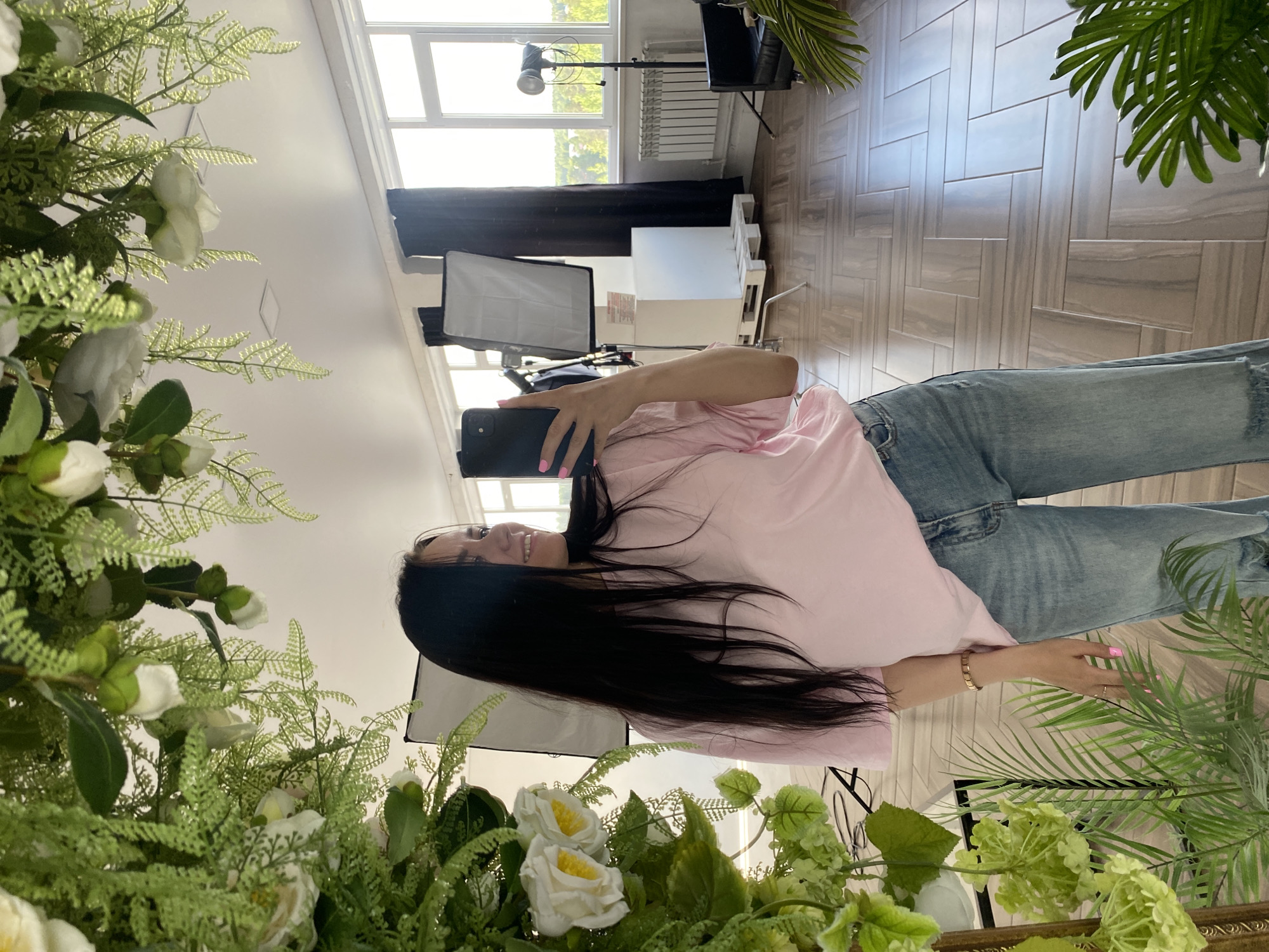 The image, rotated 90 degrees clockwise, shows a woman taking a selfie in what appears to be a photography studio in Chaikovsky, Russia. The woman, with long dark hair, is positioned in the center, facing the right side of the frame. She wears a pink short-sleeved t-shirt and light blue distressed jeans. A broad smile is visible on her face. She holds a black smartphone in her left hand, which is adorned with pink nail polish and a gold bracelet.
The foreground is framed by abundant artificial greenery and white flowers, including roses or camellias, ferns, and hydrangeas, suggesting the photo is taken through a decorative arrangement.
The background reveals a well-lit studio space. Large windows on the left side of the frame allow bright natural light into the room, with a white radiator below them and a dark curtain. Professional photography equipment, such as a large softbox on a stand and another light stand, are clearly visible. Other studio elements include a black armchair or sofa and a white low platform. The floor is covered with light-colored, patterned tiles or wood planks, arranged in a herringbone style. Green palm-like leaves from a potted plant are visible in the upper and lower right corners, contributing to the decor. The bright light through the windows suggests it is daytime. There is no visible text in the image.