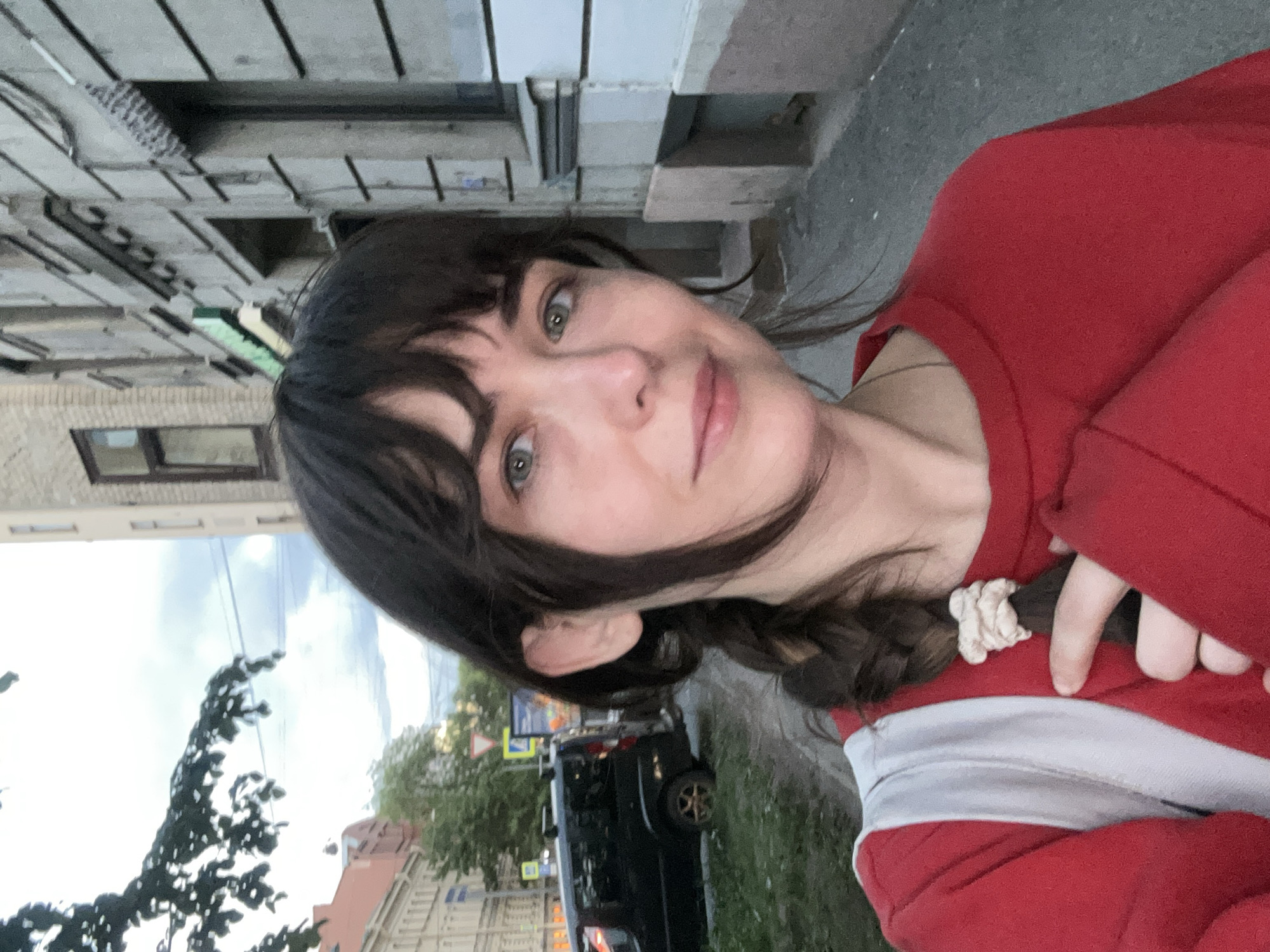 The image shows a selfie of a young woman, rotated 90 degrees clockwise. She has dark, shoulder-length hair with bangs, expressive green eyes, and a soft, gentle smile. She is wearing a red long-sleeved top featuring a prominent white horizontal stripe across the chest and stomach area. A light-colored, possibly white or cream, scrunchie holds a section of her hair in a braid.
The setting is an urban outdoor environment, consistent with Saint Petersburg, Russia. The background reveals buildings with varied architectural styles, including a section of brick and a textured facade, under an overcast or cloudy sky. Green foliage from trees is visible, along with a dark-colored SUV parked on the street. Several road signs are present, including a blue rectangular sign and a red-bordered warning triangle, though any text or specific symbols on them are illegible. The lighting is diffused, suggesting an overcast day or possibly late afternoon. No specific activity beyond posing for the selfie is depicted.